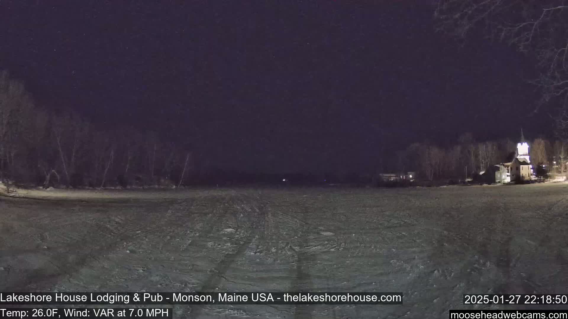 A nighttime view of a snow-covered frozen lake with bare trees and a building in the distance under a dark sky.