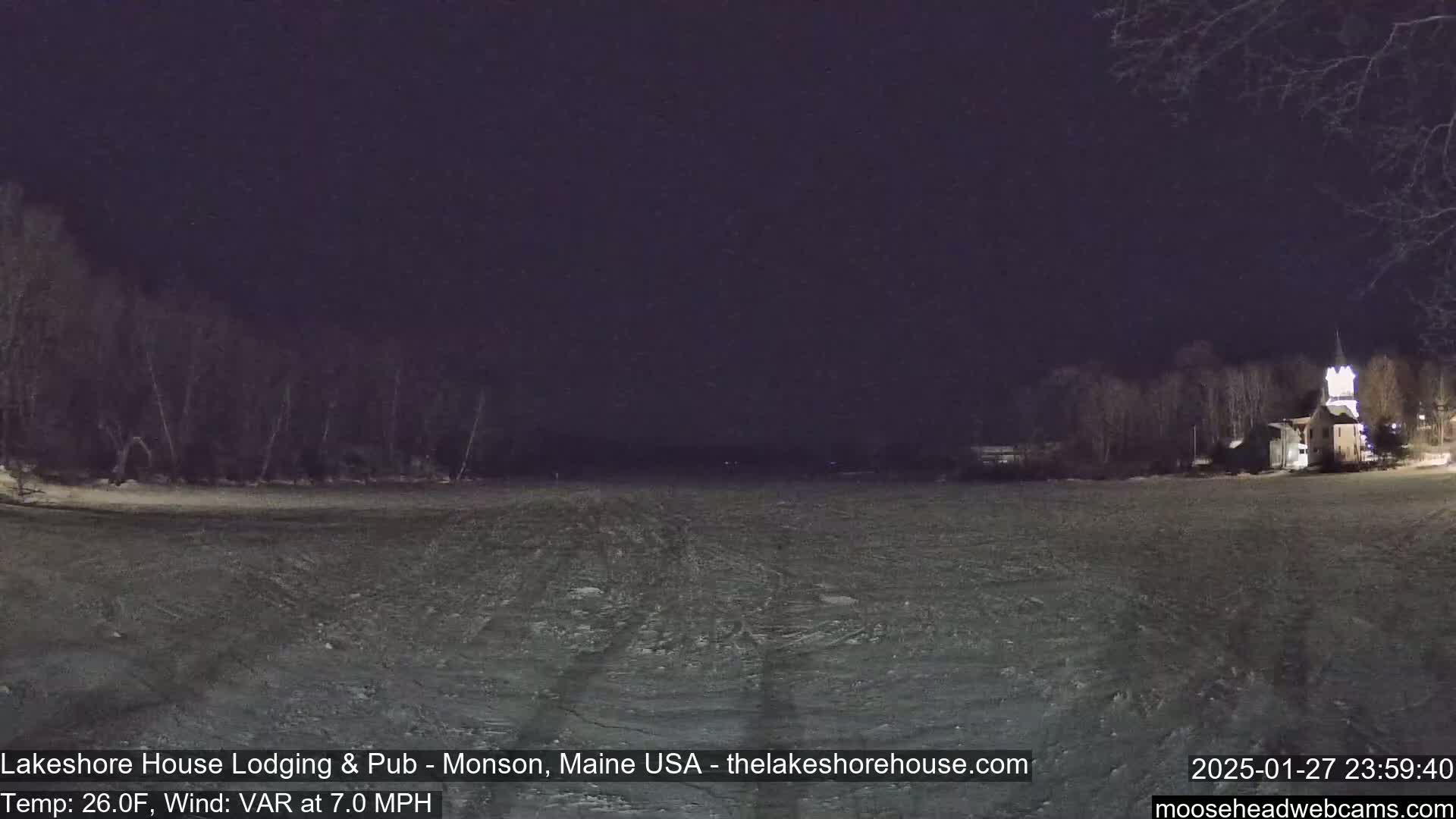 A nighttime, snow-covered field is visible, with dark trees and a building with lights in the background under a dark sky, and a 7 mph wind.