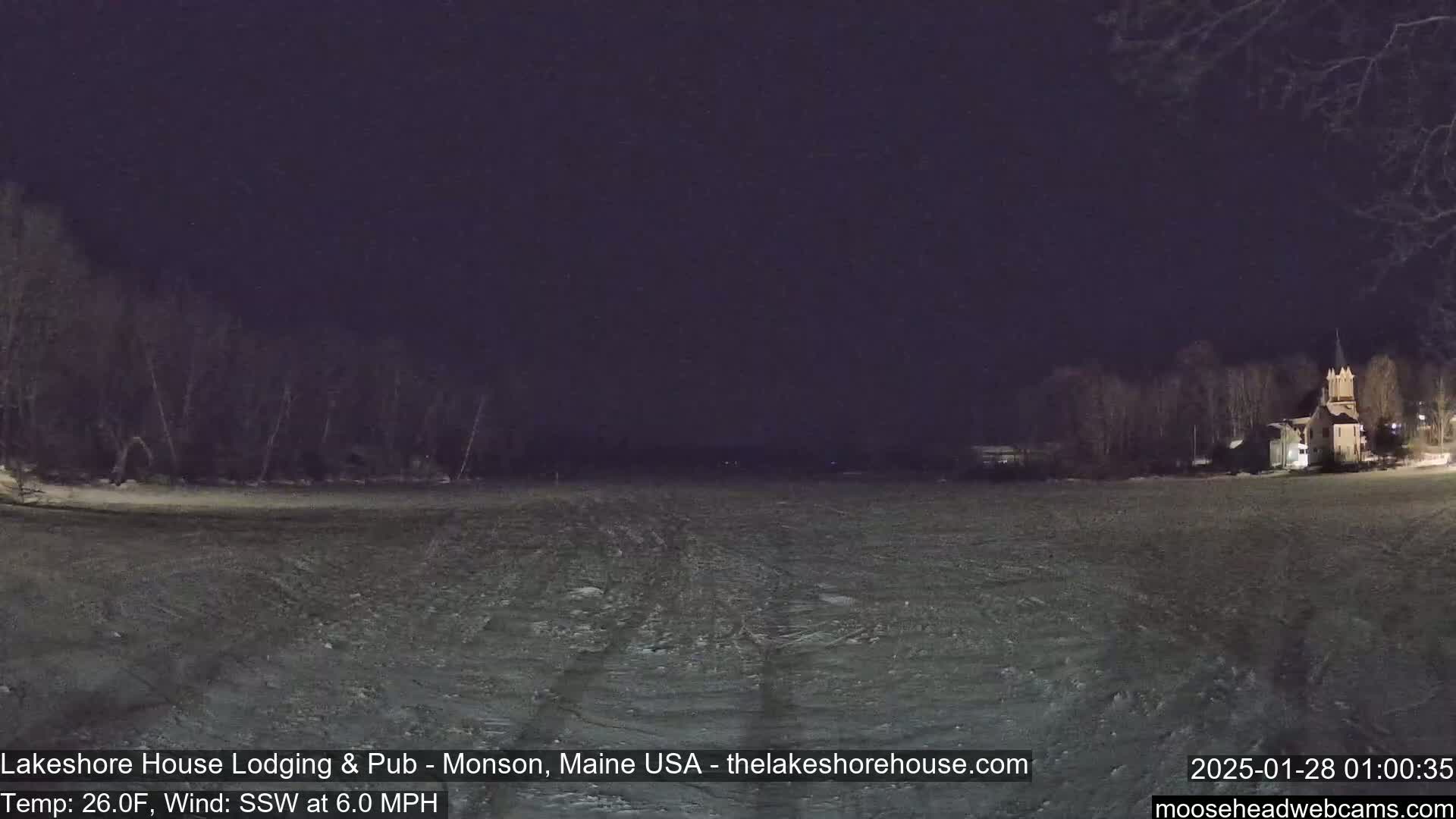 A snow-covered field at night, under a dark sky, shows bare trees and a building with a steeple in the distance, with a 6 mph wind blowing from the south-southwest at 26 degrees Fahrenheit.