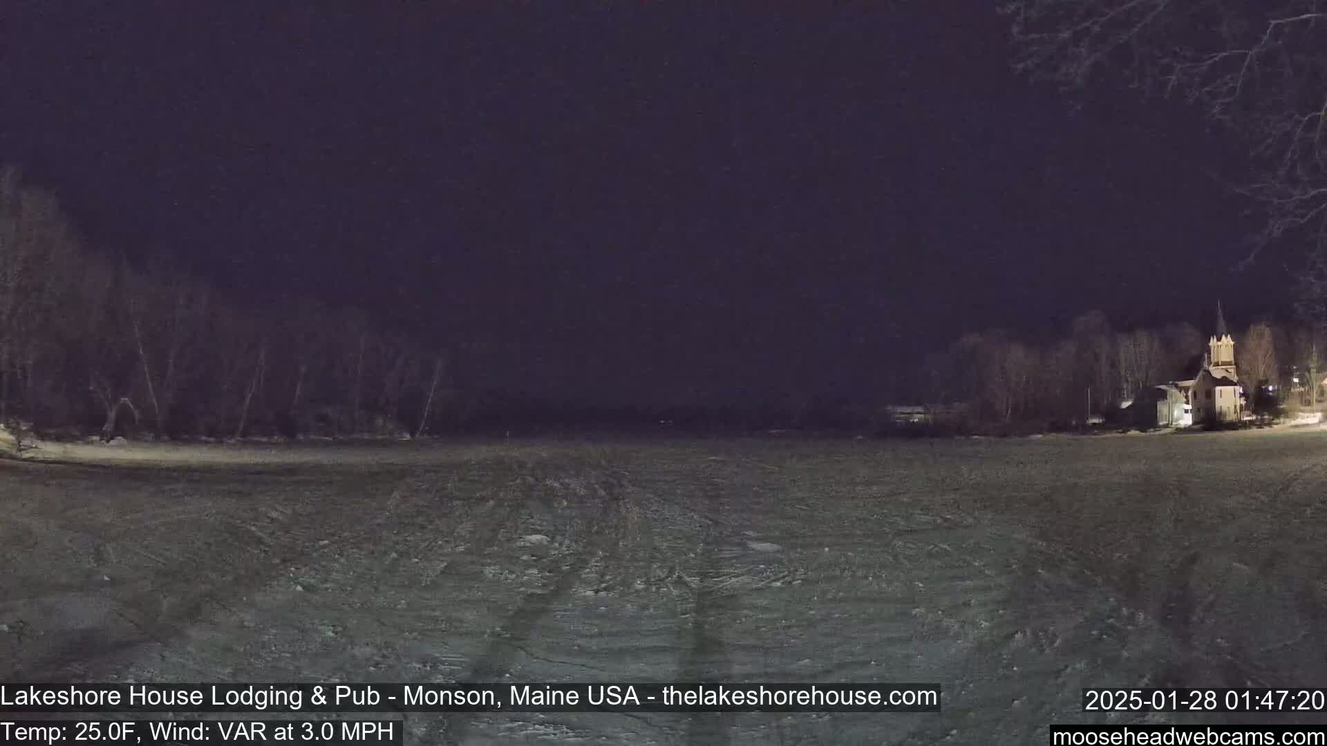 A snow-covered field at night is illuminated by a building with lights on in the distance under a dark sky.