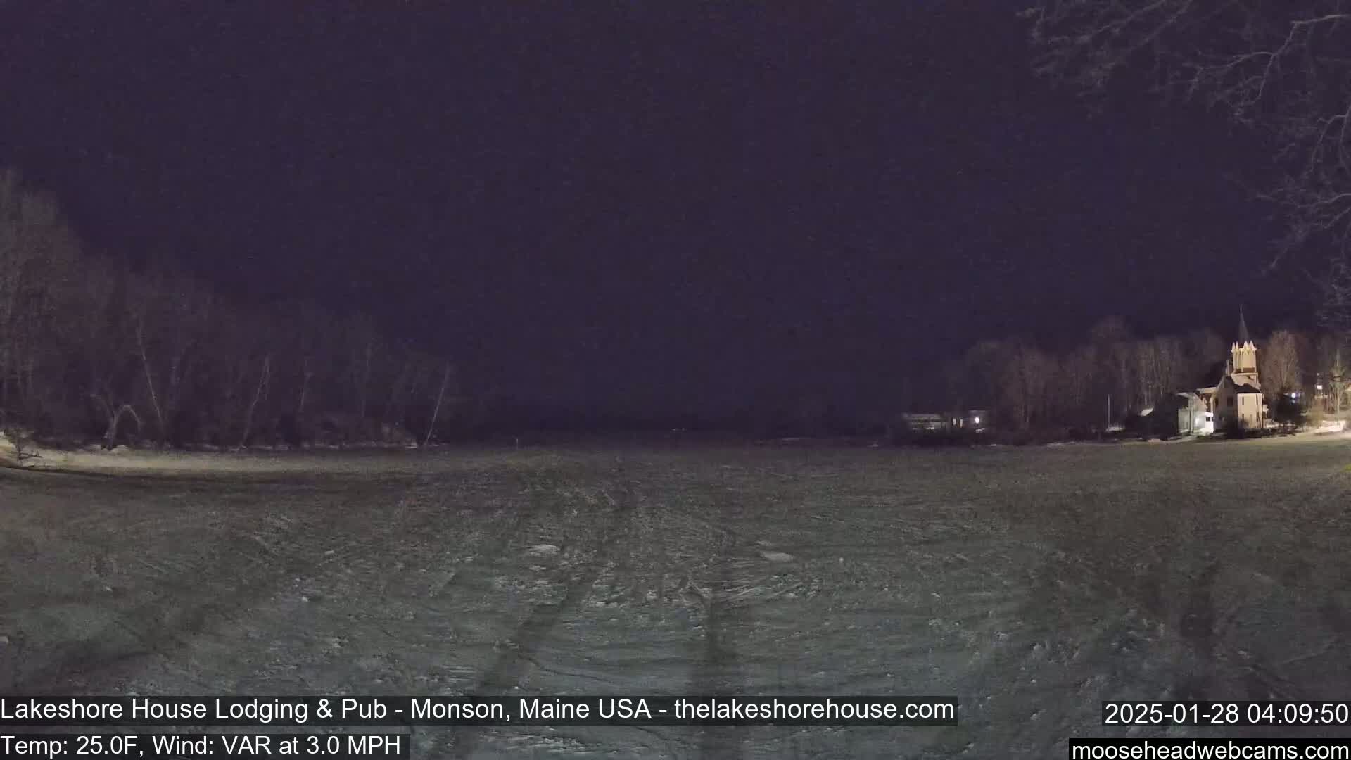 A nighttime view of a snow-covered field, with dark trees and a building with a steeple in the background under a dark sky.
