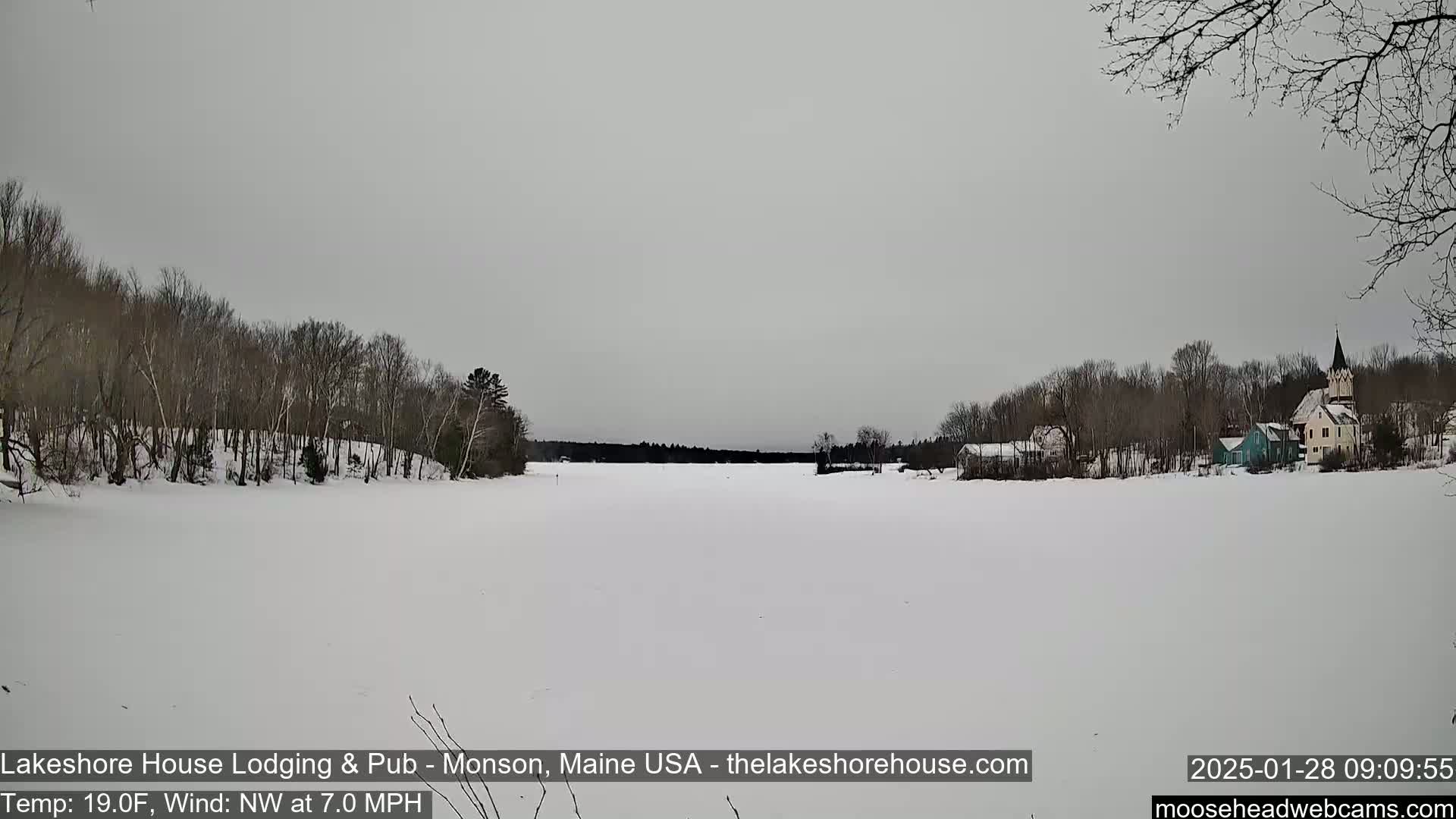 A frozen lake is seen with a village of houses and a church steeple on the far shore, under an overcast sky.
