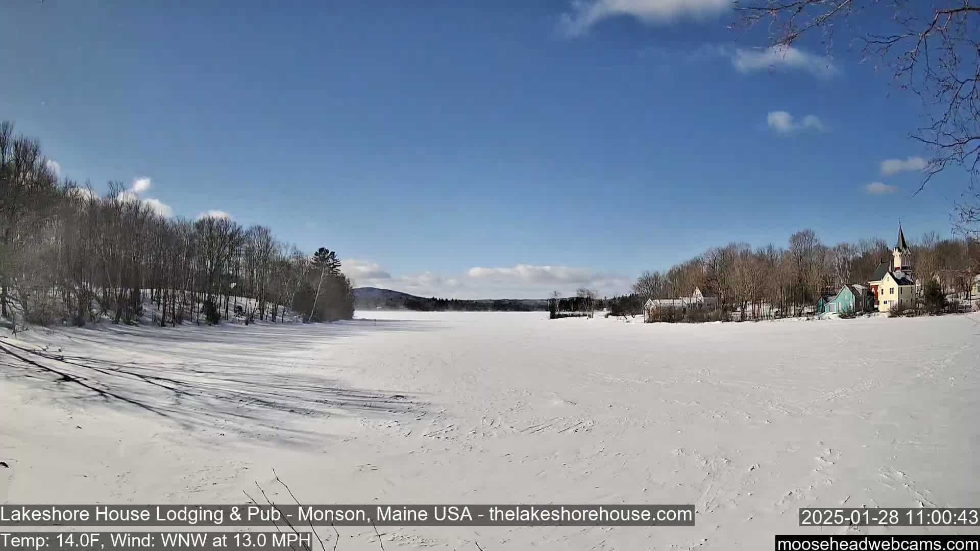 A snow-covered frozen lake is seen under a mostly sunny sky, with a small town and bare trees visible in the background.