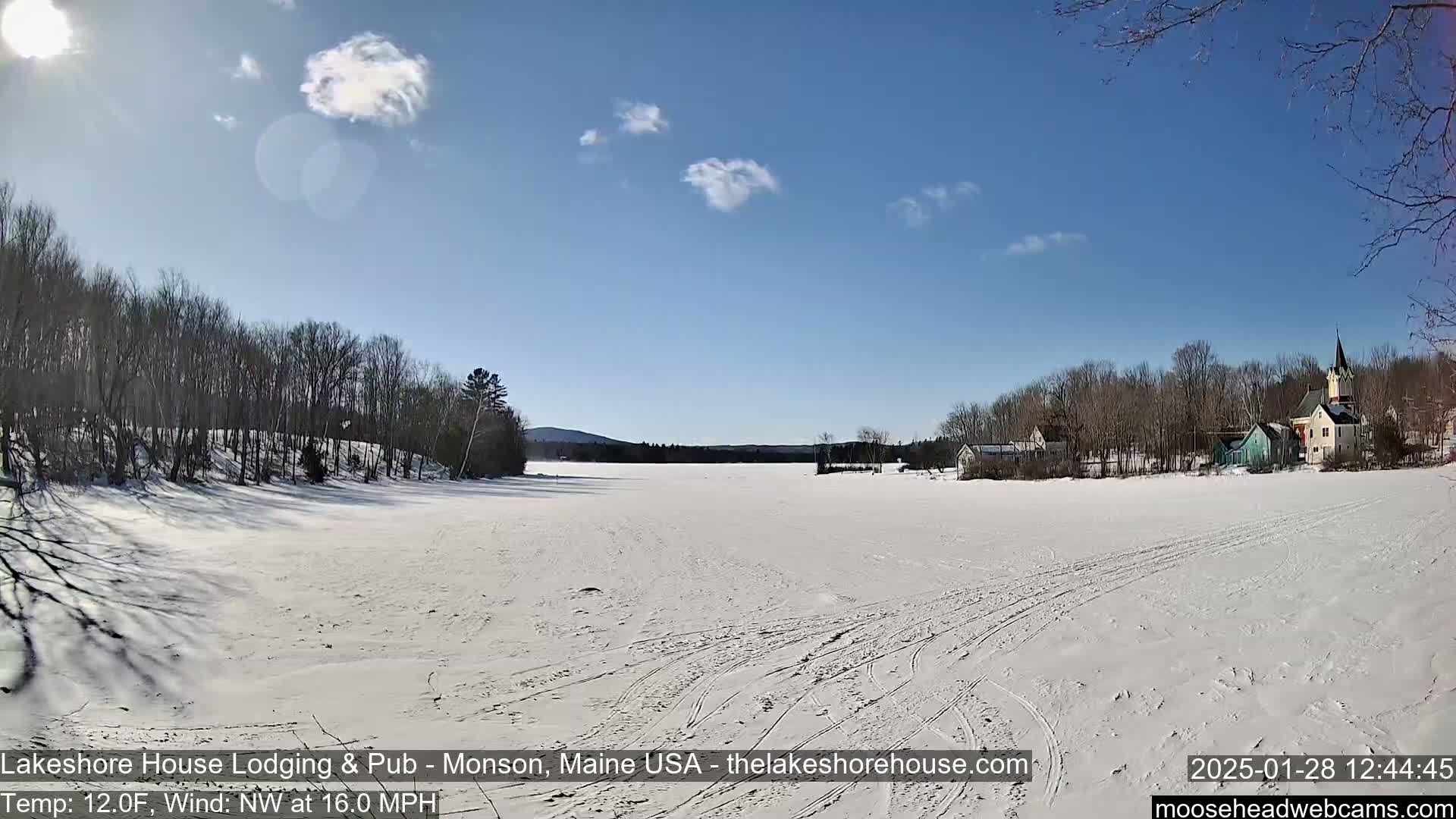 A snow-covered frozen lake stretches out to a cluster of buildings and trees under a mostly sunny sky.