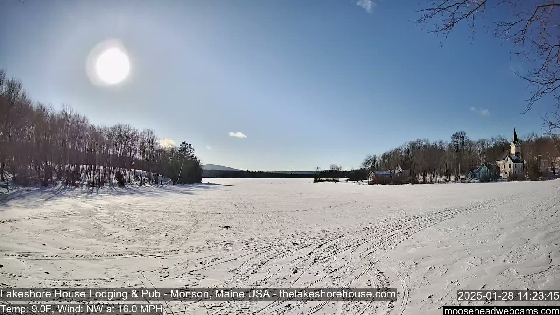 A frozen lake stretches out under a bright, sunny sky, with snow-covered trees and buildings in the background, and a brisk northwest wind.