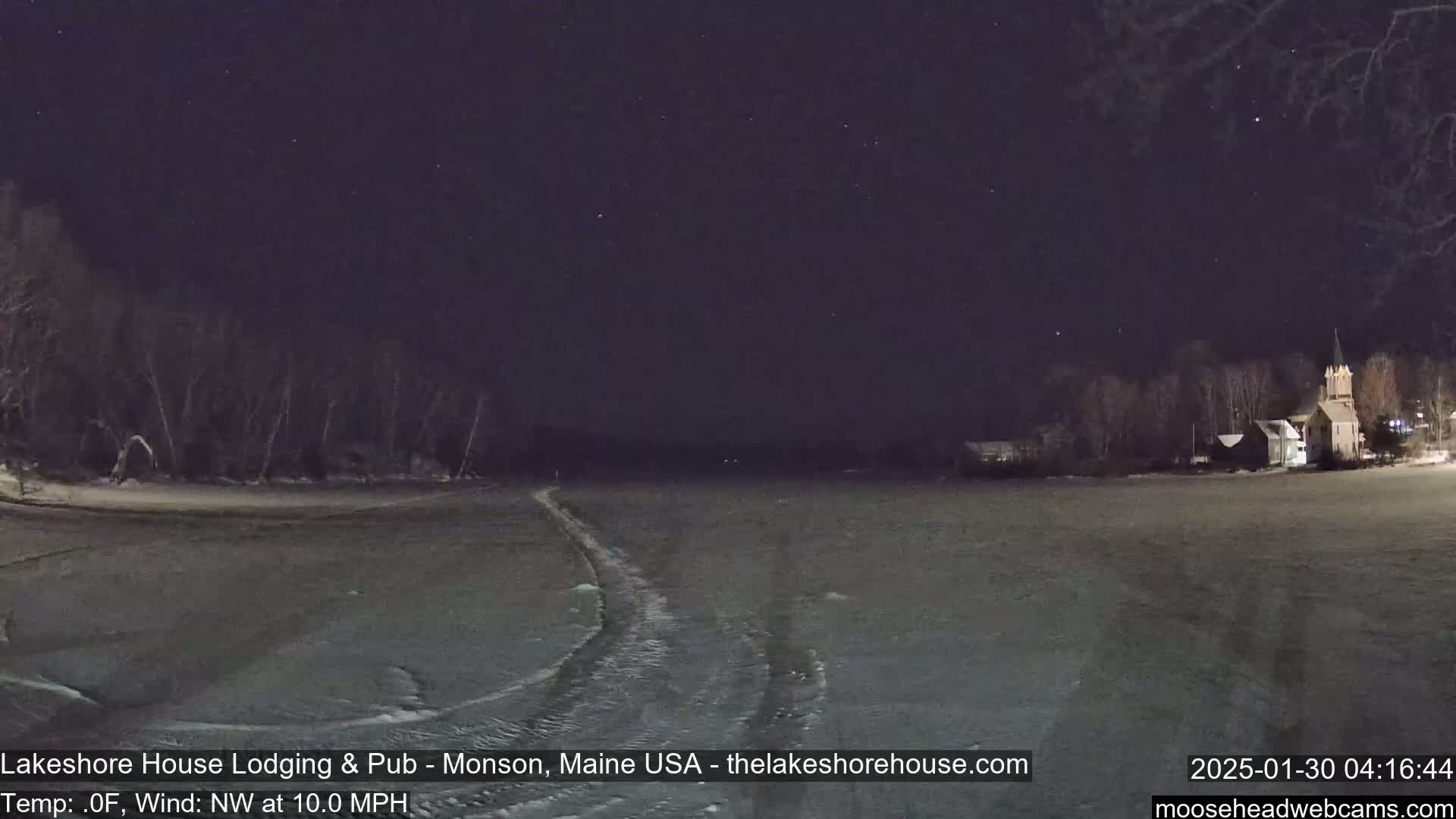 A snow-covered field at night under a starry sky, with a building visible in the distance and tire tracks in the foreground, with a northwest wind at 10 mph and a temperature of 0°F.