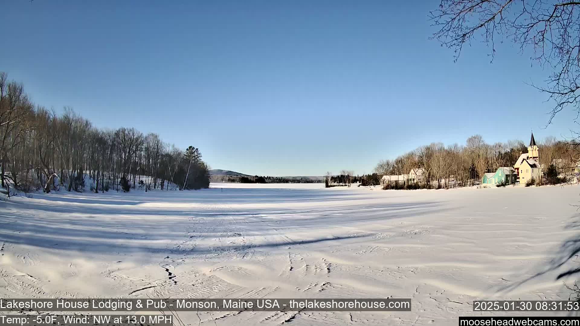 A frozen lake, under a clear blue sky, is surrounded by snow-covered trees and a few buildings.