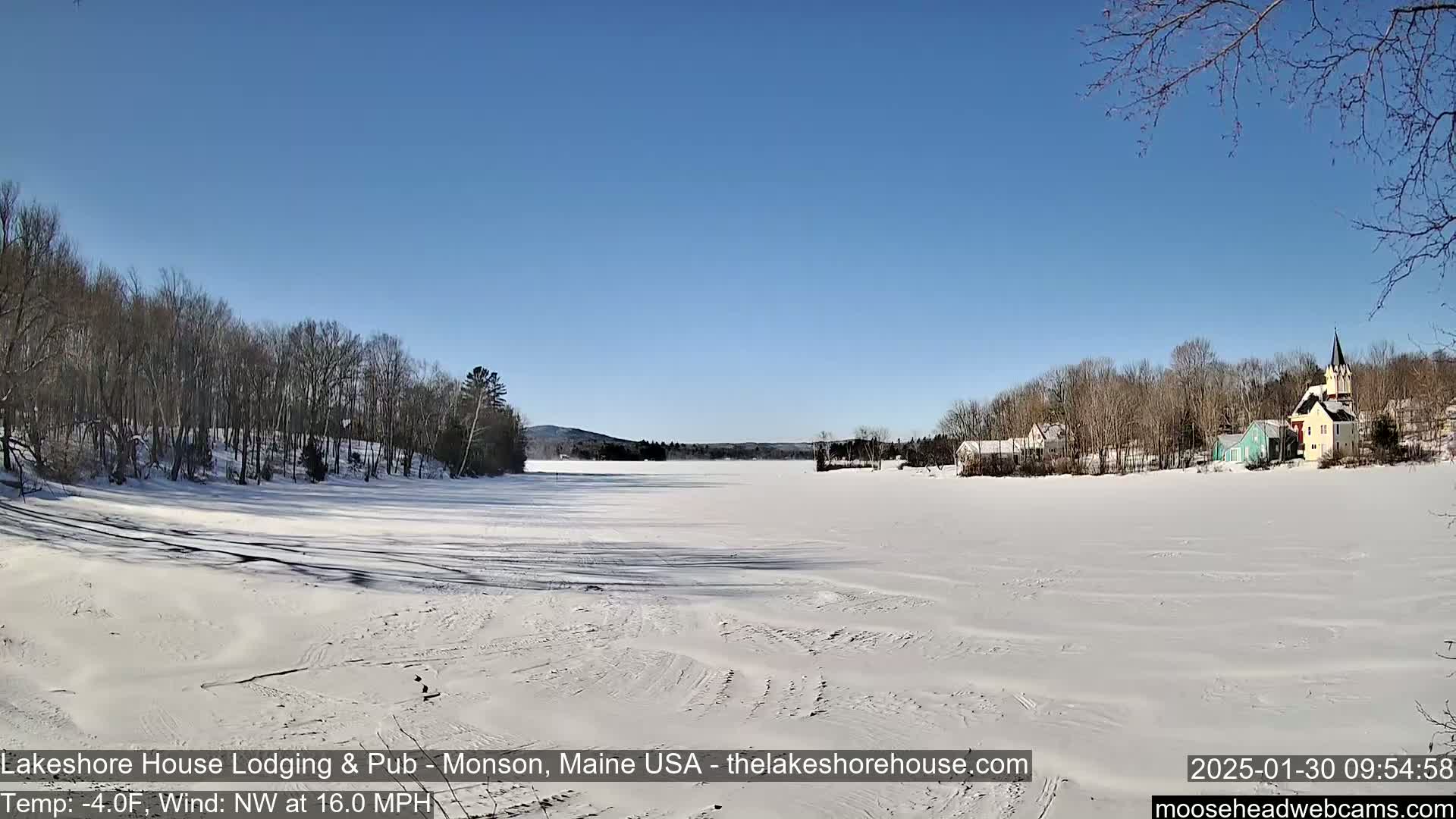 A frozen lake stretches out under a clear blue sky, with snow-covered trees and buildings lining its far shore.