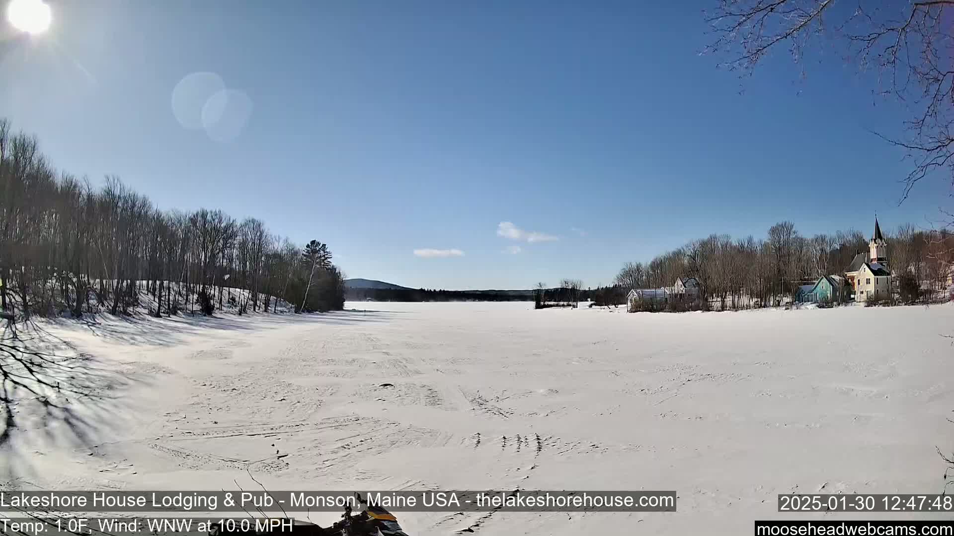 A snow-covered frozen lake stretches out to a small town of buildings and trees under a bright, sunny sky.