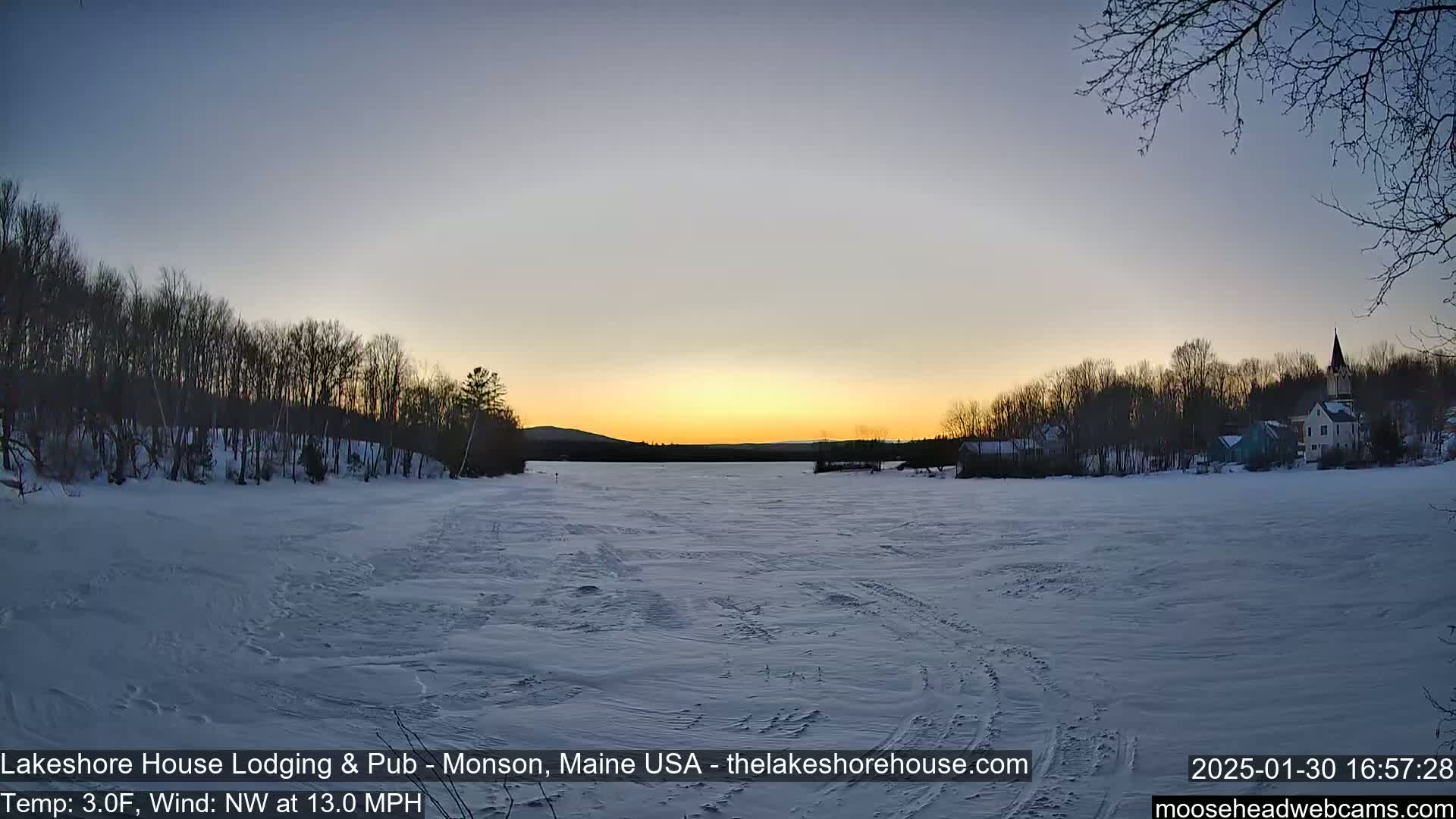 A frozen lake at sunset is surrounded by snow-covered trees and a small town in the distance, under a clear sky with a northwest wind at 13 mph and a temperature of 3°F.