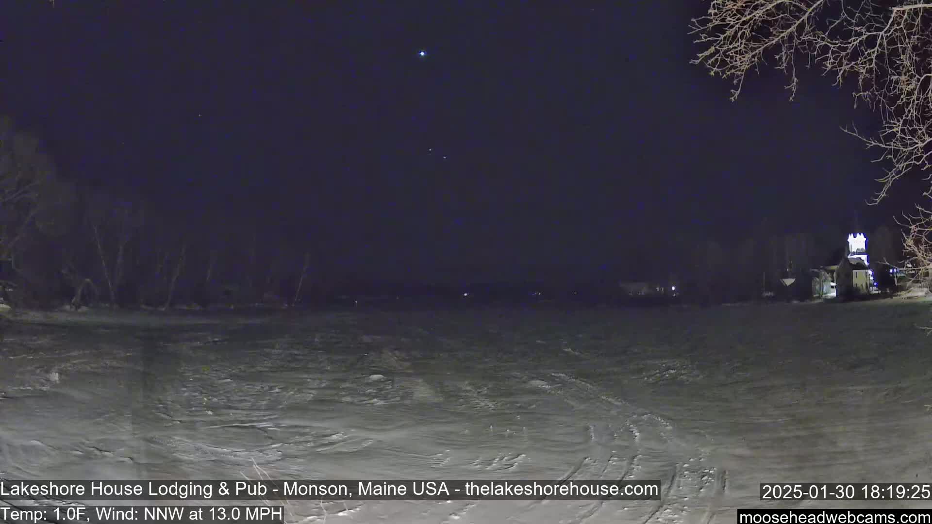 A nighttime view of a snow-covered expanse, under a dark sky with a few visible stars, with some buildings and bare trees in the distance; a 13 mph wind blows from the north-northwest at a temperature of 1°F.