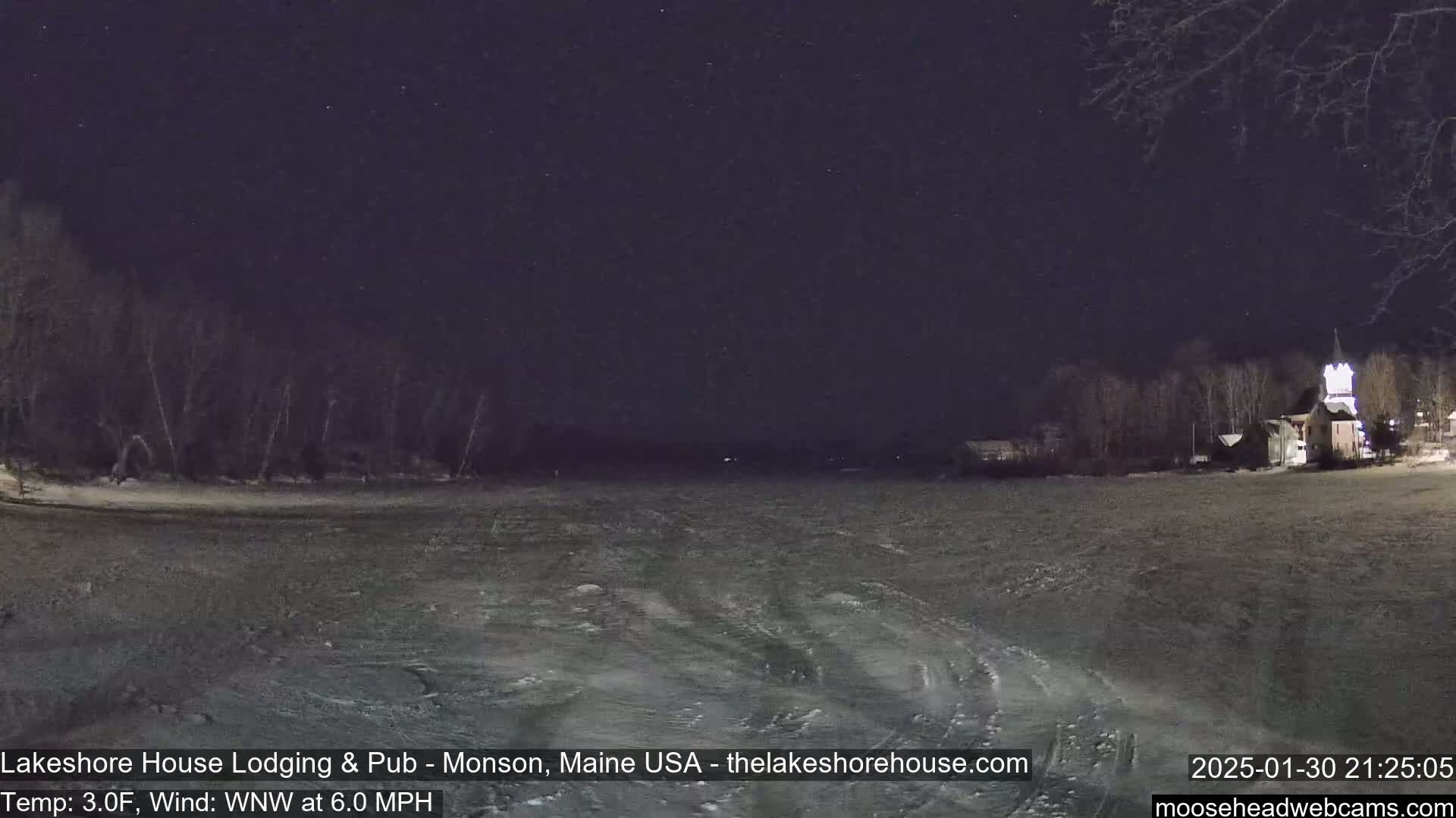 A nighttime view of a frozen lake under a clear, starry sky, with bare trees and a building visible on the far shore.