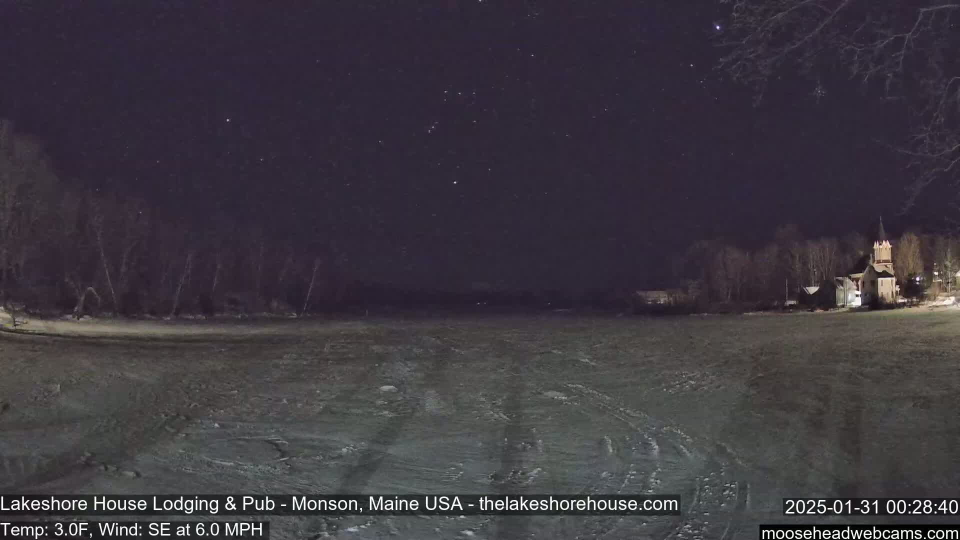 A snow-covered field at night under a starry sky, with a few buildings and trees in the distance.