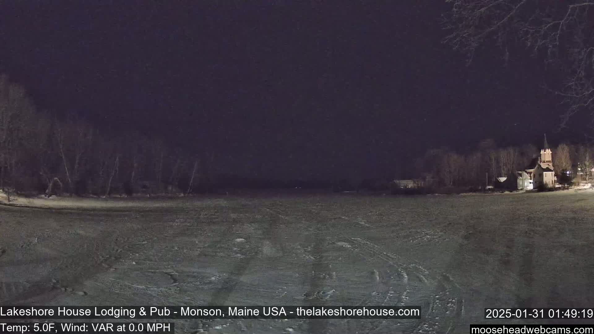 A dark, nighttime scene shows a frozen body of water with tire tracks, bordered by snow-covered land and trees, and a building with a steeple in the distance.