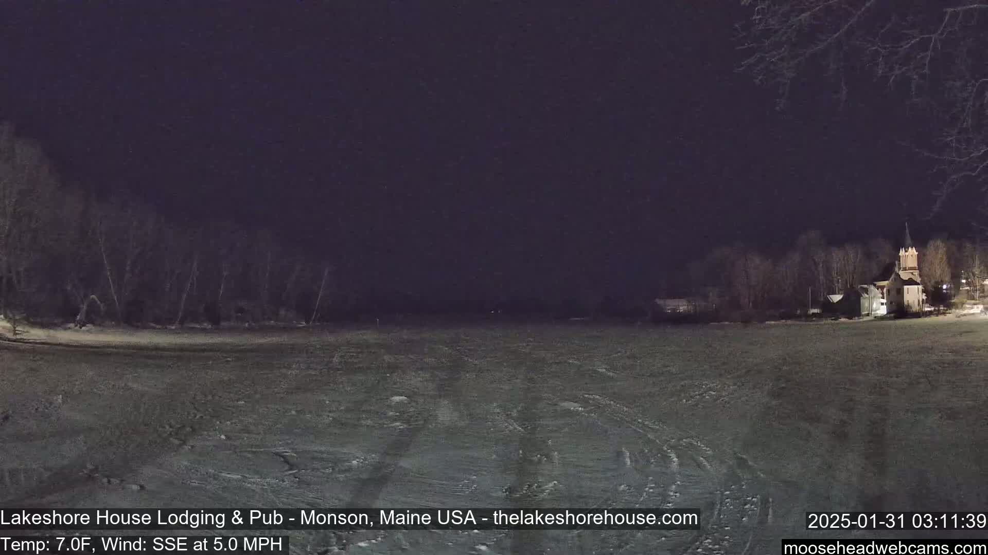 A snow-covered field at night, under a dark sky, shows a building with a steeple in the distance and trees along the shoreline.