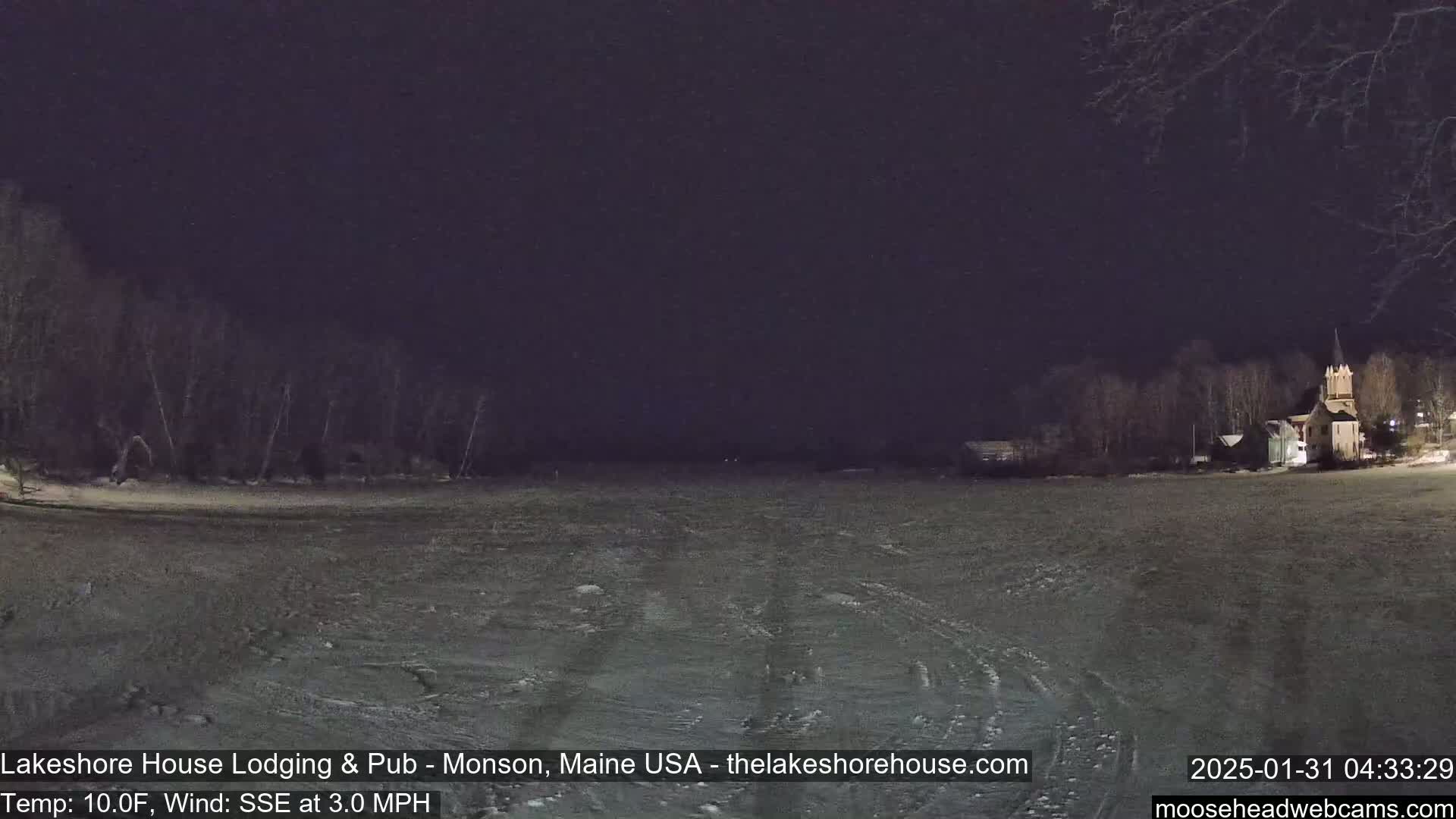 A snow-covered, flat expanse stretches to a dark, tree-lined shore at night, with a building faintly visible in the distance under a dark sky.