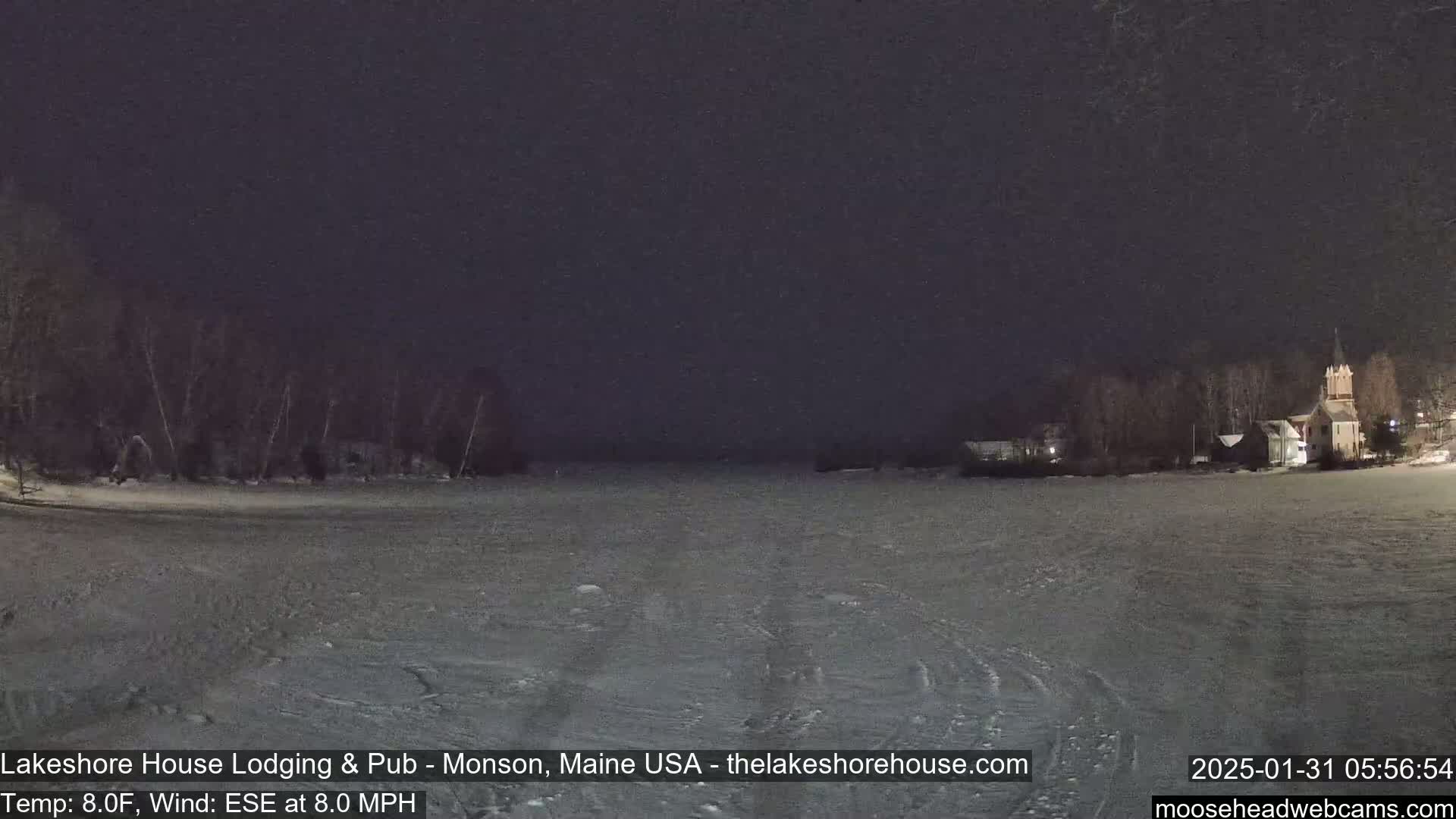 A snow-covered frozen lake at night, with dark trees and a building with a spire visible in the distance under a dark sky.