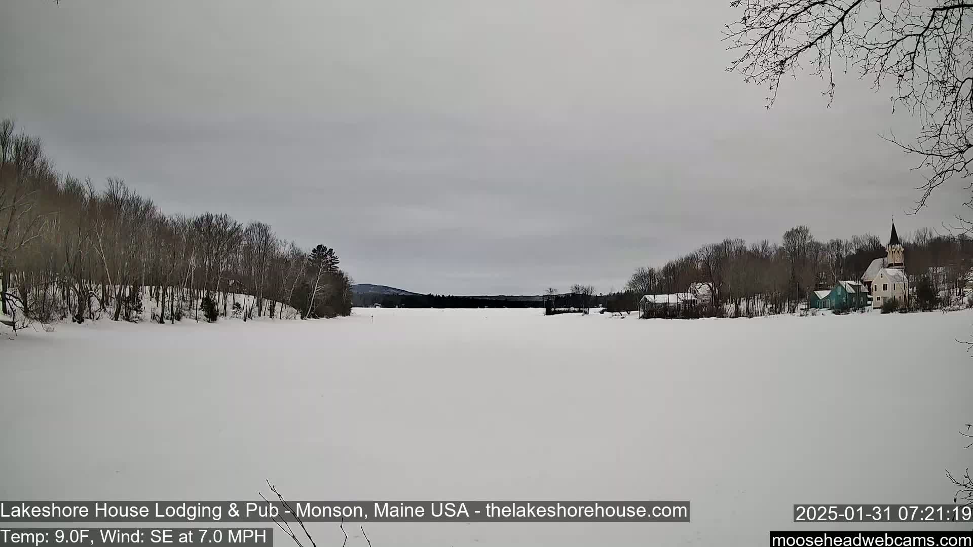 A snow-covered lake stretches out to a small village with a church steeple under an overcast sky.