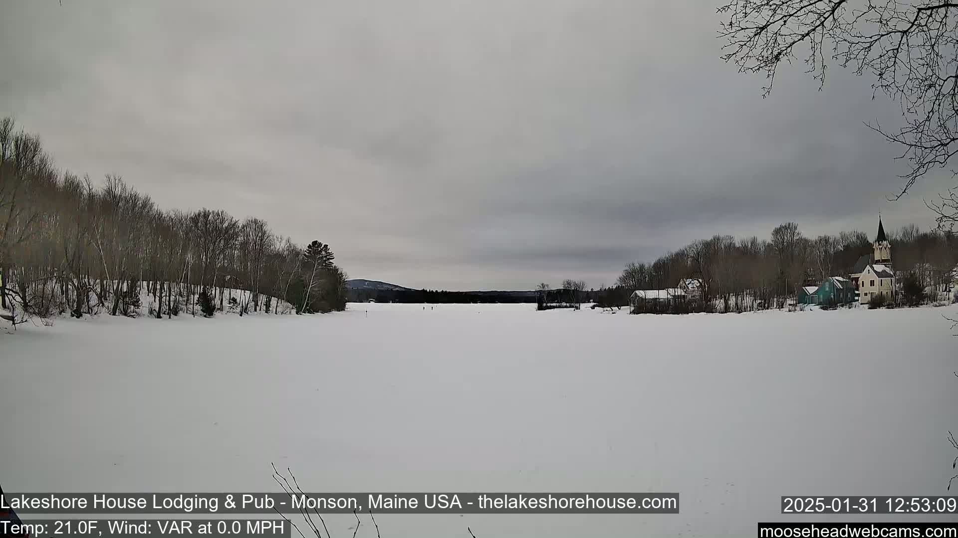 A frozen lake stretches to the far shore where a small town with a church steeple is visible under an overcast sky.