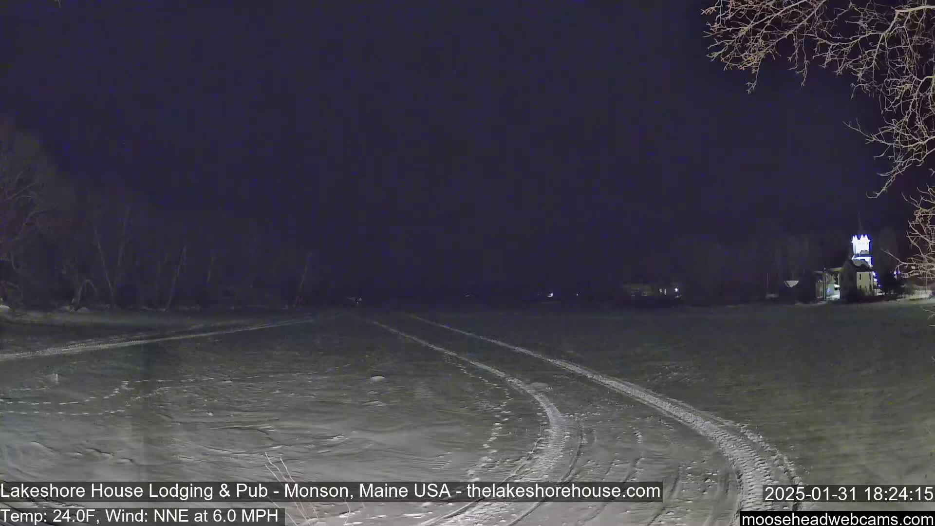 A snow-covered field at night with tire tracks leading toward a building in the distance, under a dark sky with a NNE wind at 6.0 mph and a temperature of 24°F.