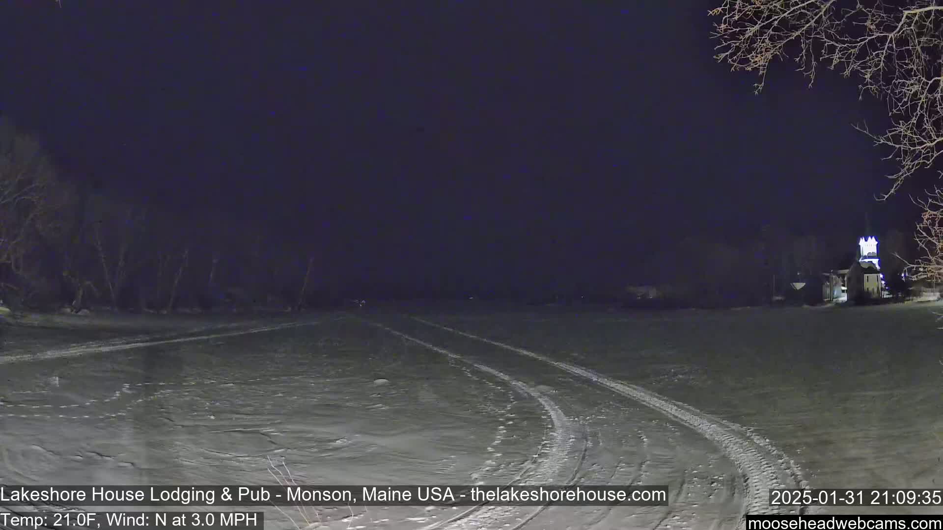 A snow-covered field at night with tire tracks, a building in the distance, and bare trees under a dark sky.