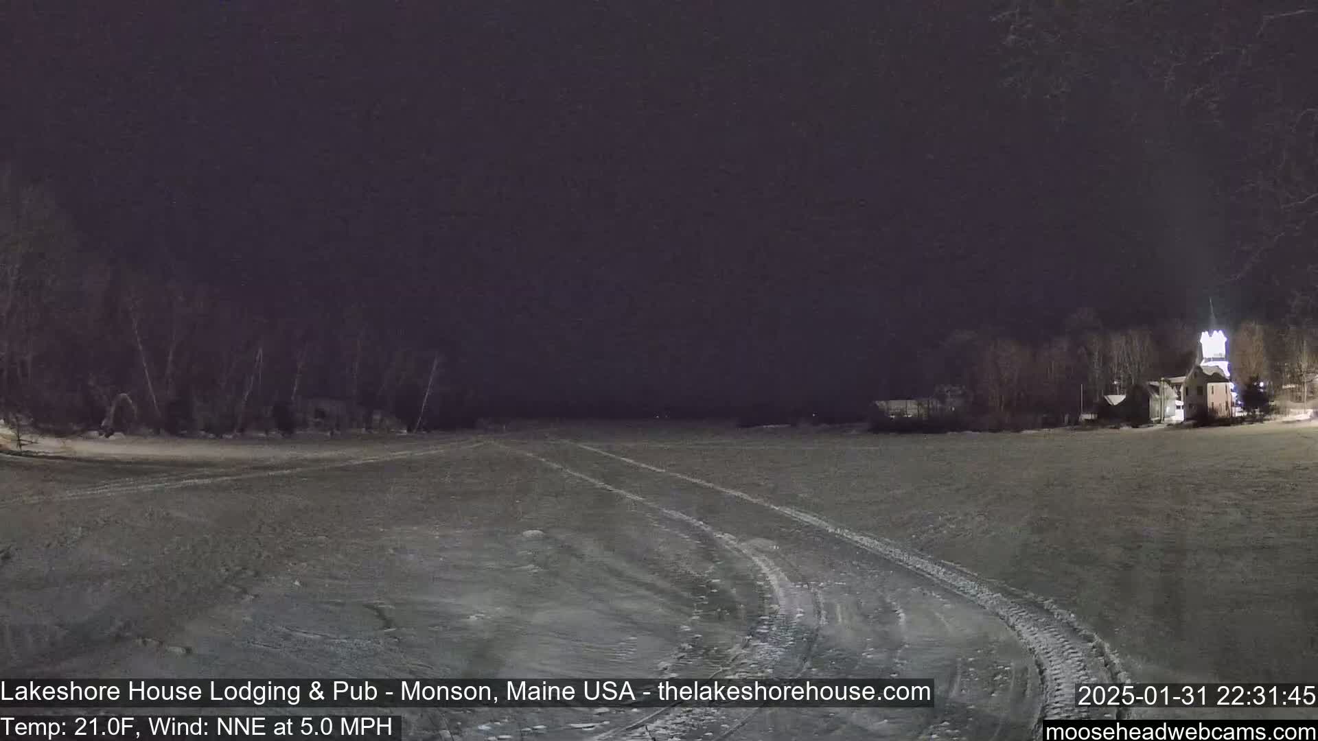 A snow-covered, flat expanse is visible at night, with tire tracks winding through it toward a dimly lit building in the distance under a dark sky.