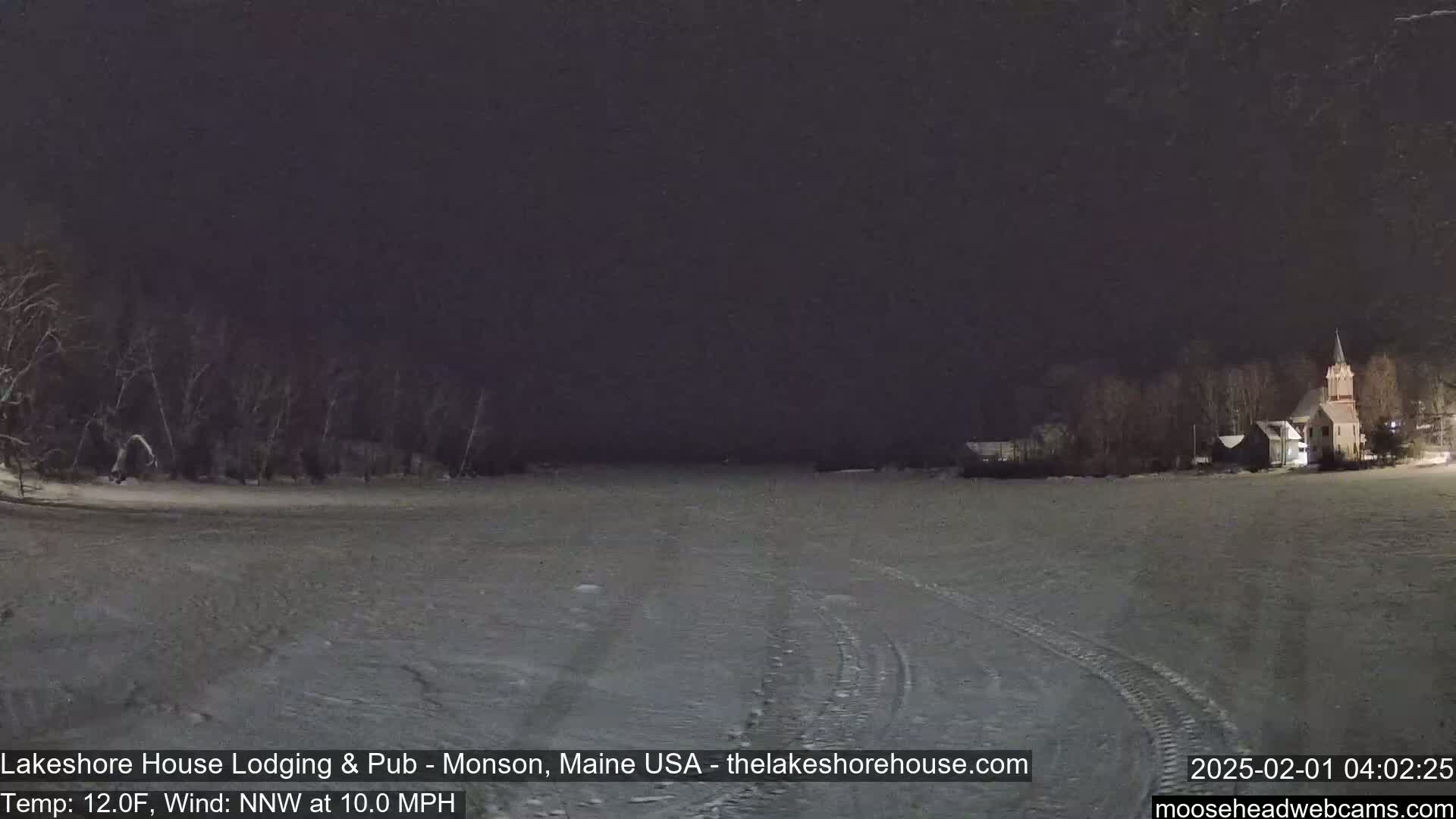 A snow-covered field at night, with a church and trees visible in the distance under a dark sky, with a 12°F temperature and a 10 mph northwest wind.
