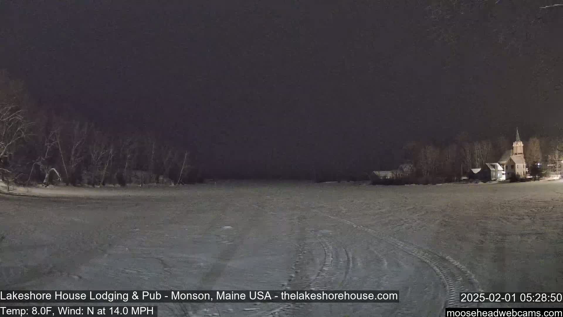 A snow-covered expanse of land stretches out under a dark night sky, with a building visible in the distance, and trees lining the left side of the image.