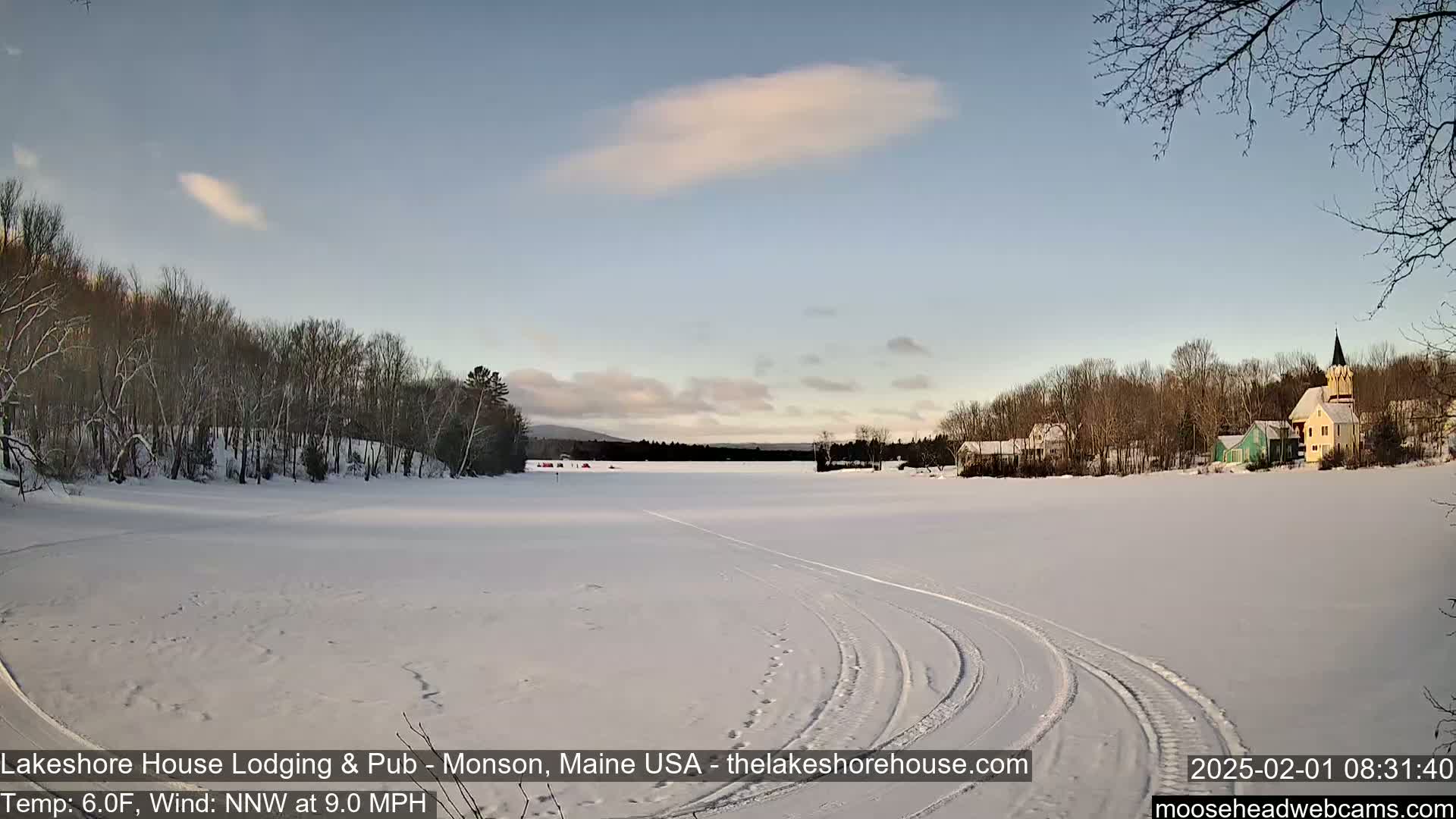 A snow-covered frozen lake with tire tracks, bare trees lining the shore, and a few buildings visible in the distance under a mostly clear, light blue sky.