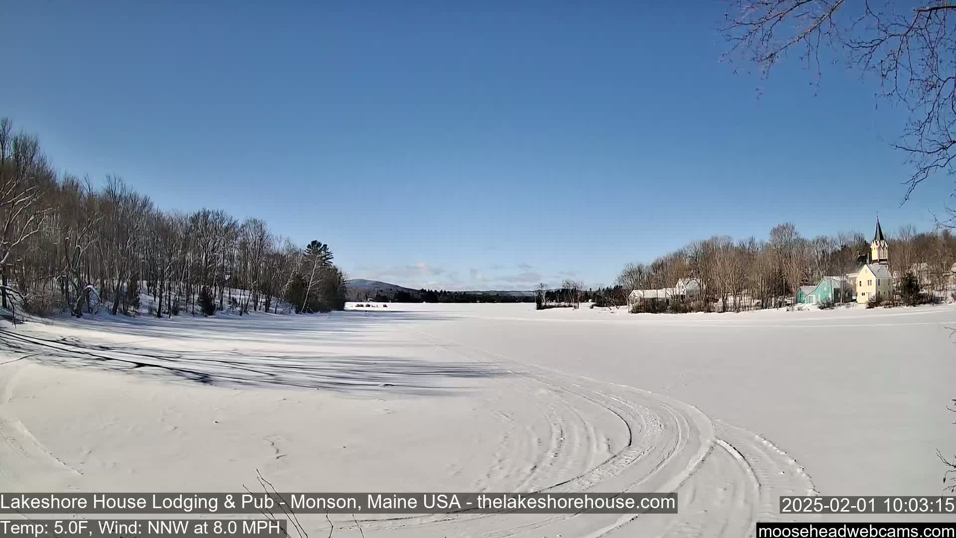 A snow-covered frozen lake stretches towards a small village of houses and a church under a clear blue sky, with a light wind blowing.