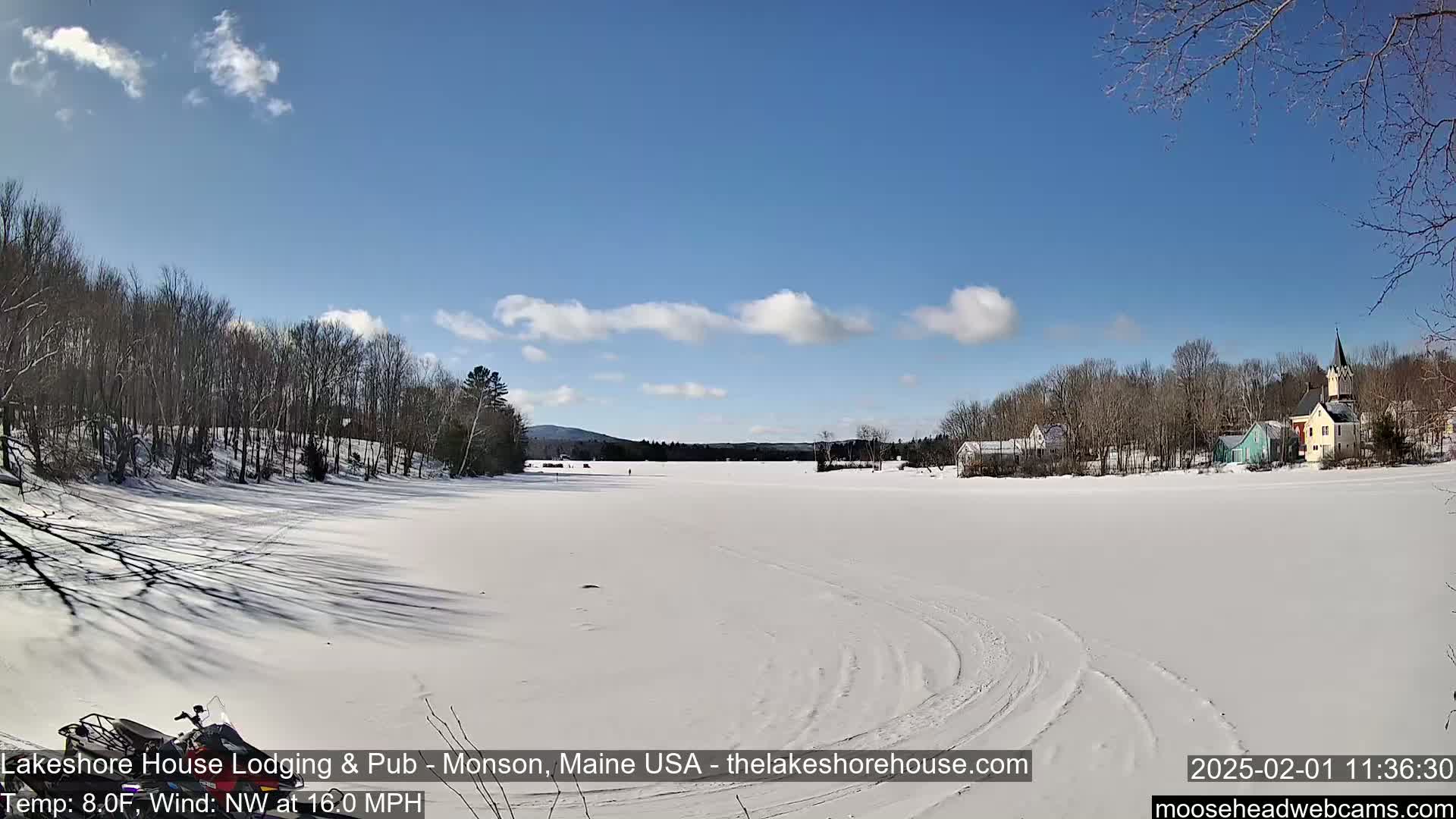 A snow-covered frozen lake stretches toward a small village under a mostly clear, bright blue sky.