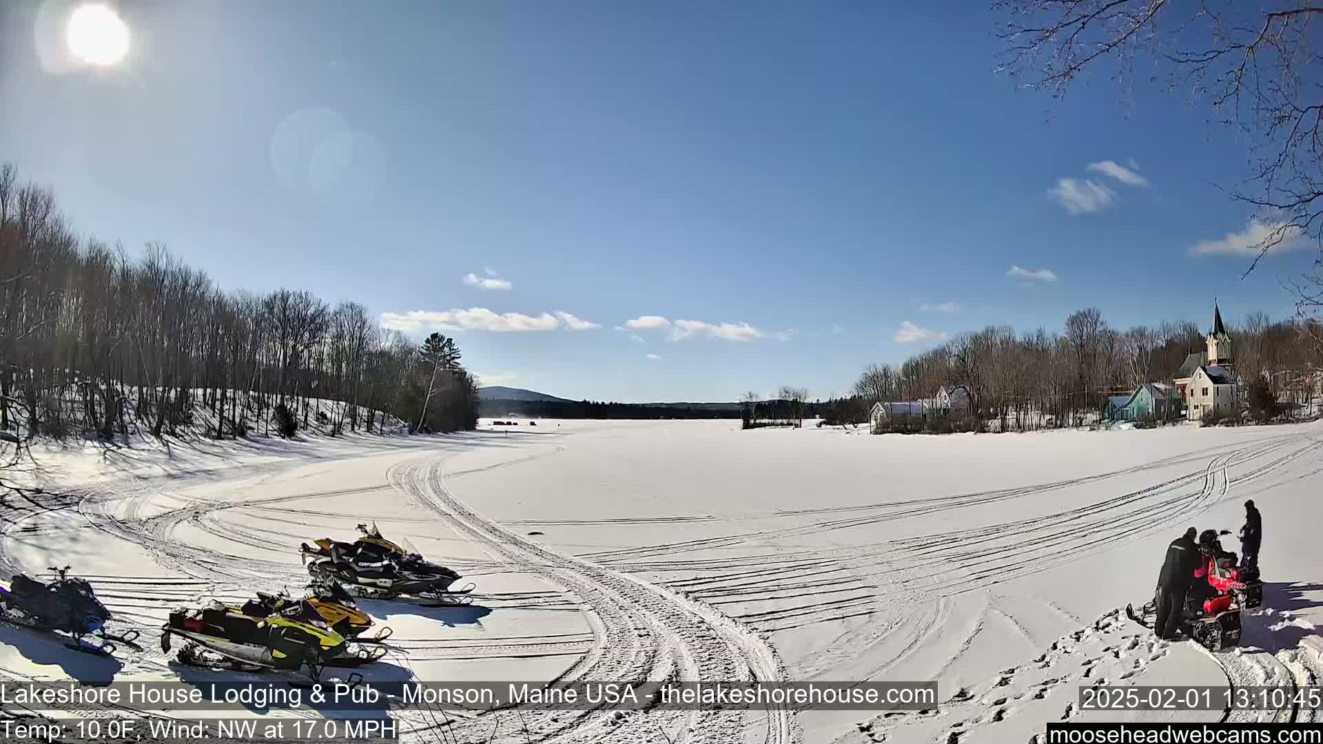 A snow-covered frozen lake is seen on a sunny day, with snowmobiles parked near the shore and a few people near others, across from a small village of houses and trees.