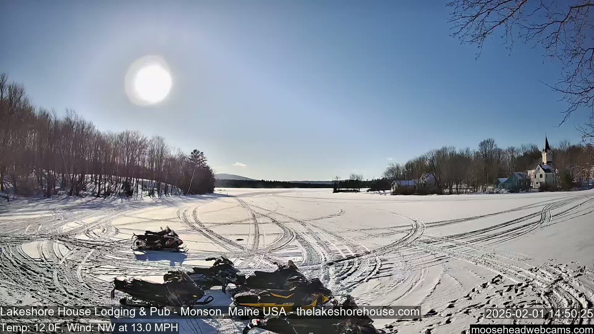 Several snowmobiles are parked on a snow-covered frozen lake under a bright, sunny sky, with a village visible in the distance.