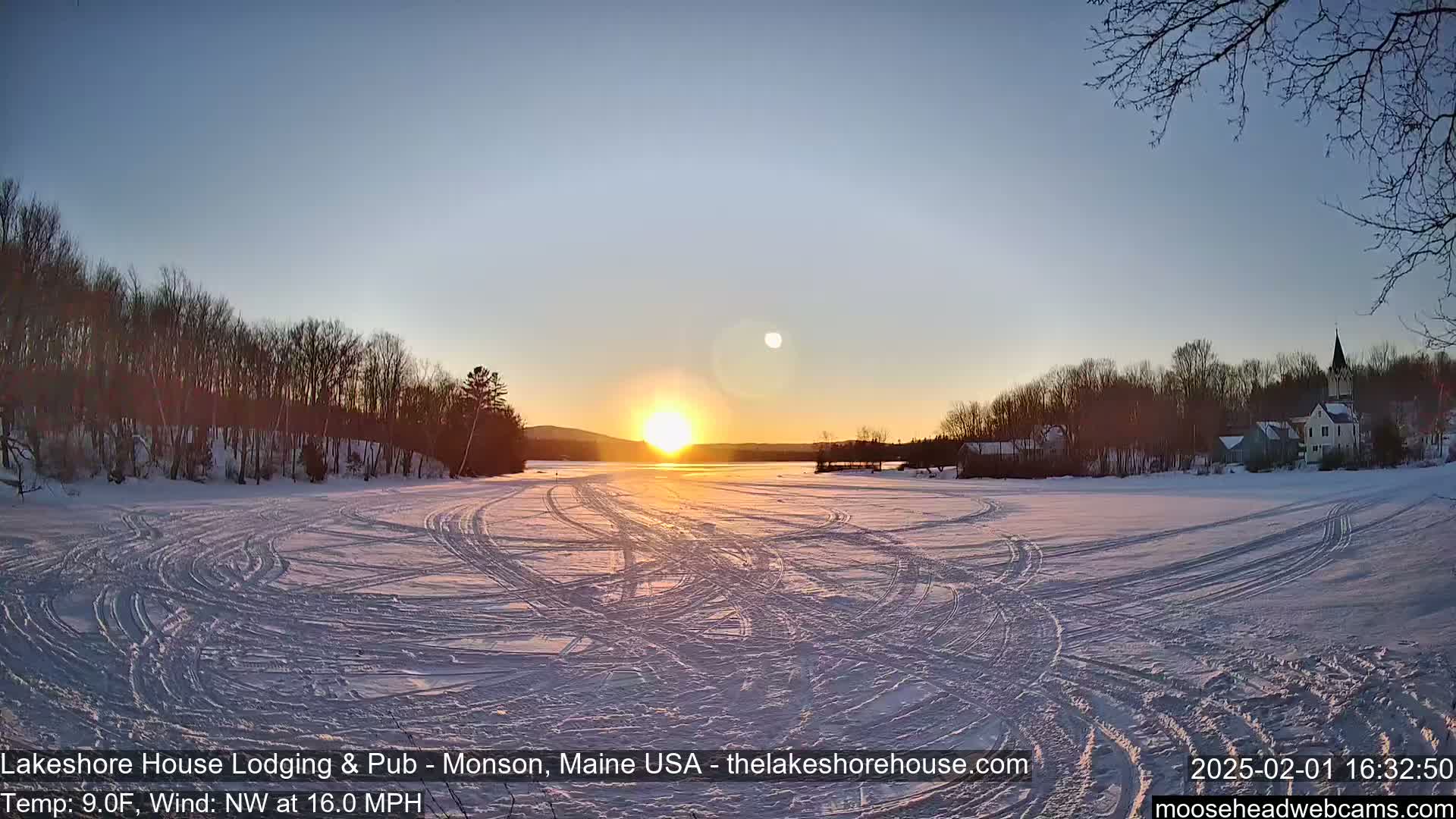 A frozen lake with tire tracks, surrounded by snow-covered trees and buildings, shows a setting sun in a cold, windy, clear sky.