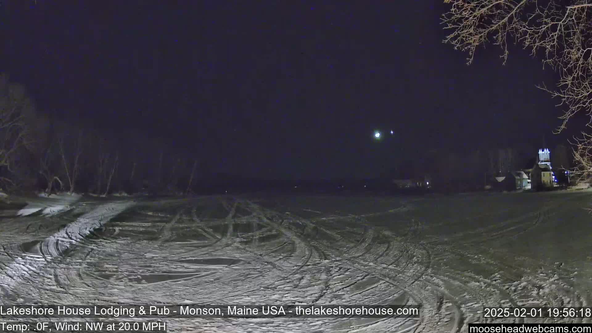 A snow-covered field with tire tracks under a dark, clear night sky, lit by a distant light source.