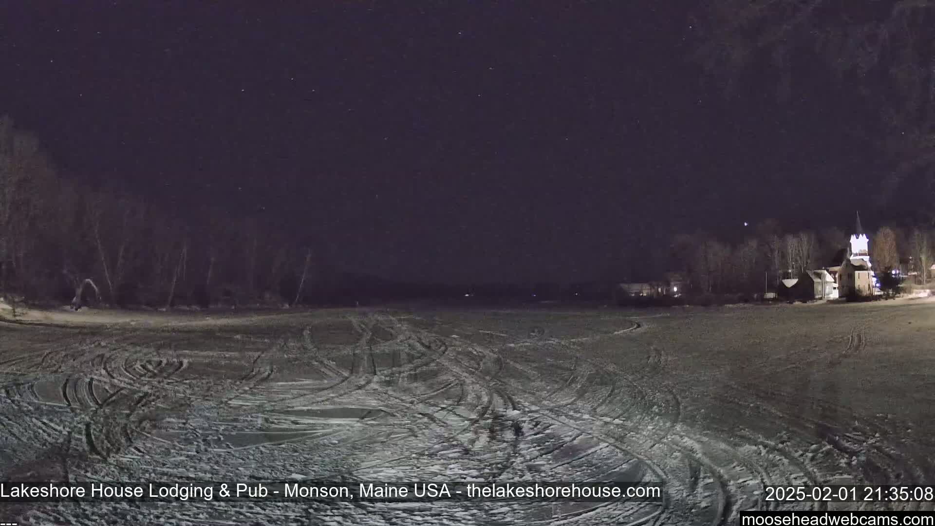 A snow-covered, track-marked expanse stretches under a clear, starlit night sky, with a line of trees and buildings visible in the distance.