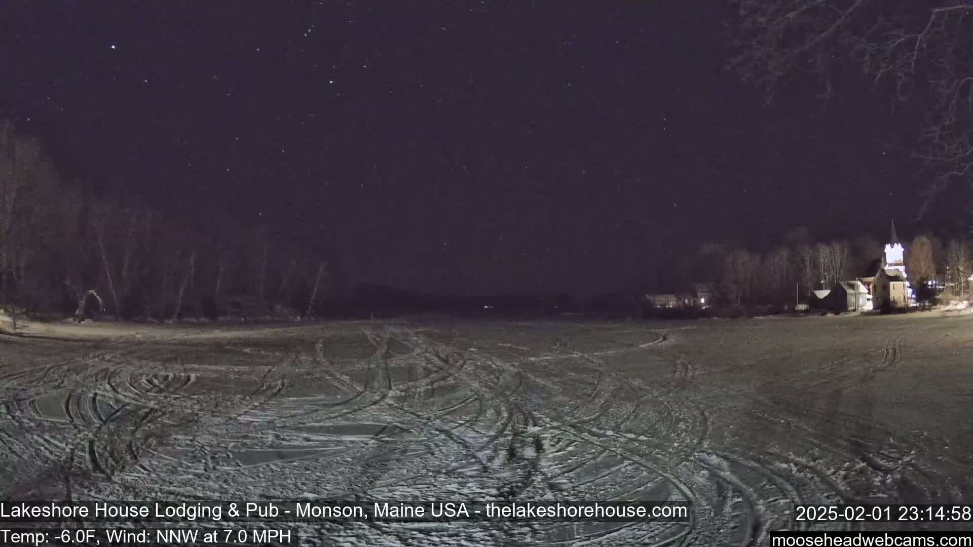 A snow-covered, flat expanse with tire tracks under a clear, starry night sky shows a few buildings in the distance.