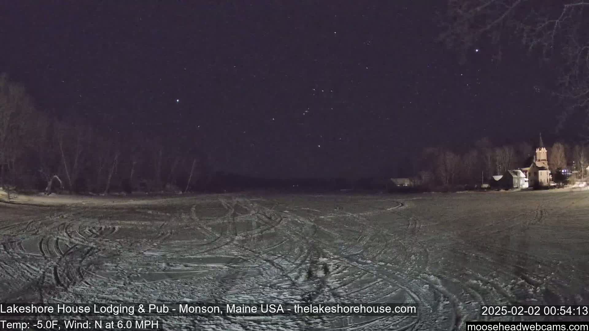 A snow-covered field with tire tracks under a starry night sky, a few buildings visible in the distance, and a north wind at 6.0 mph with a temperature of -5°F.