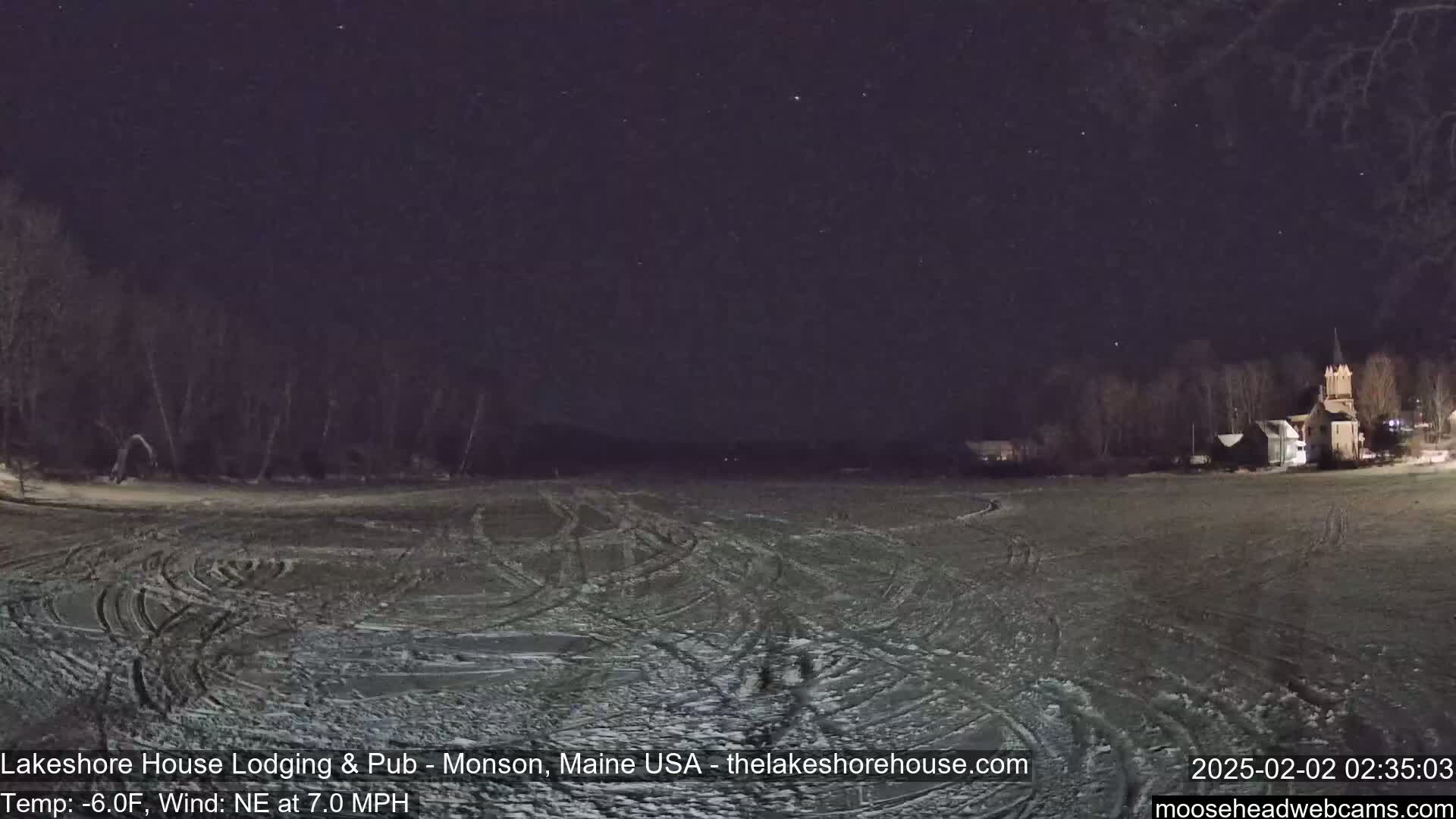 A snow-covered field with tire tracks under a clear, starry night sky, and buildings in the background.