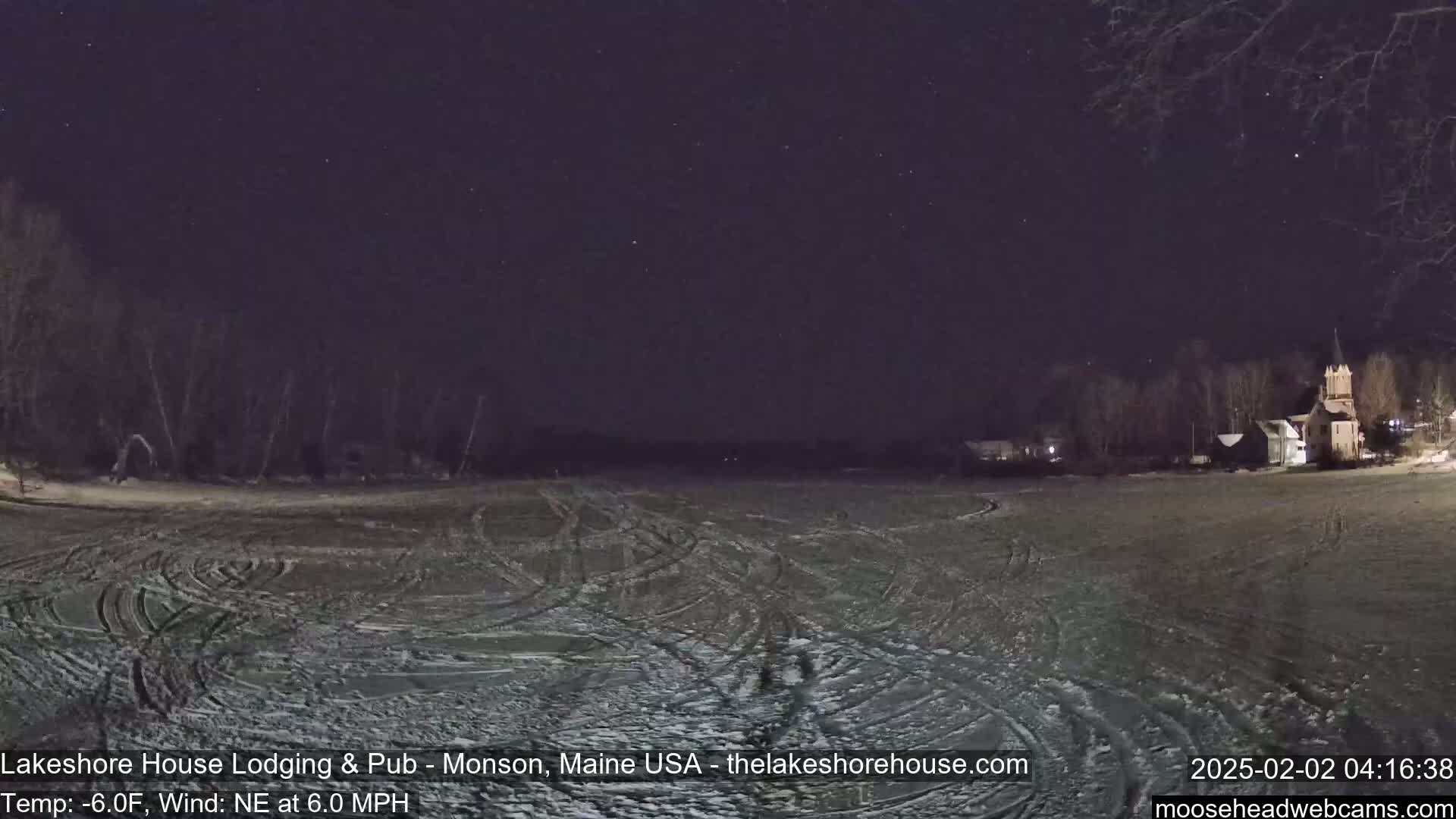 A snow-covered, frozen lake with tire tracks under a clear, starry night sky shows a few buildings in the distance.