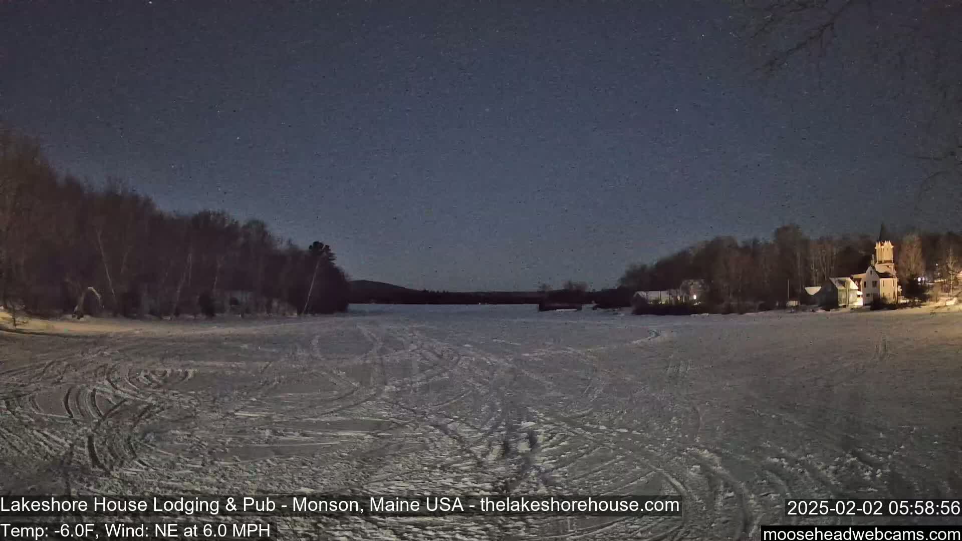 A snow-covered frozen lake at night, under a starry sky, with a shoreline of dark trees and buildings visible in the distance.