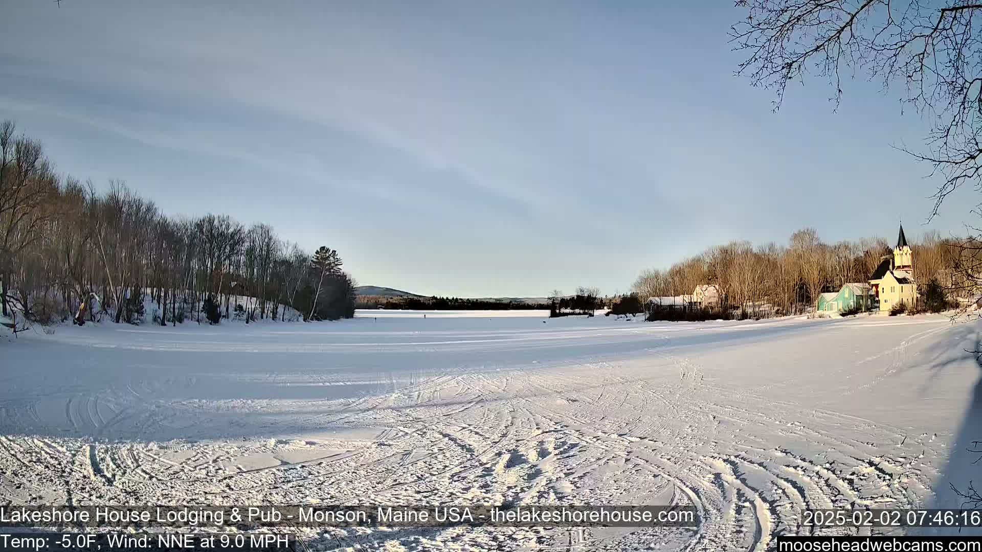 A frozen lake, crisscrossed with tracks, stretches before a line of snow-covered trees and buildings under a clear, bright sky.