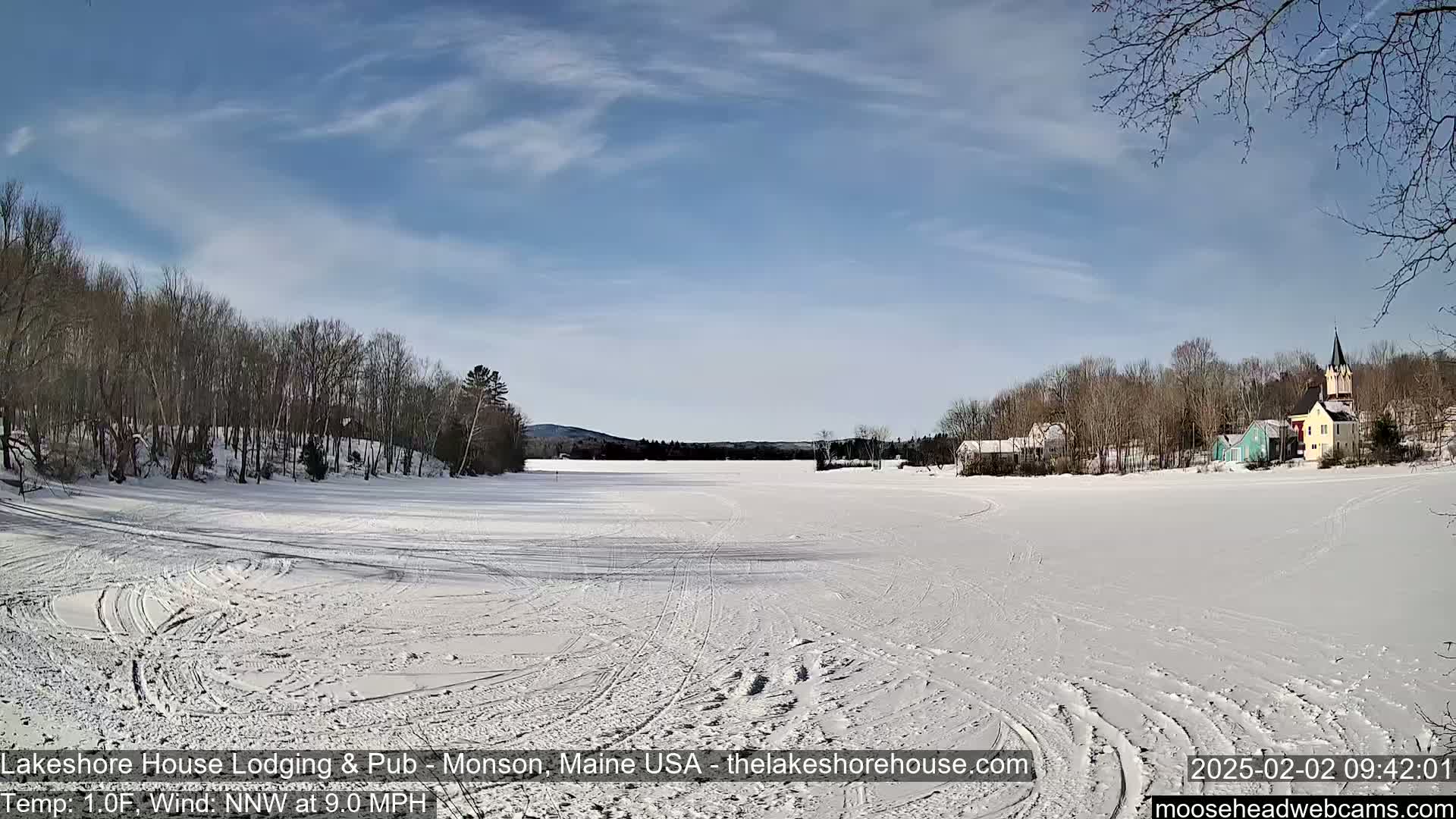 A snow-covered frozen lake stretches out before a village of houses and trees under a mostly sunny sky with a light wind.