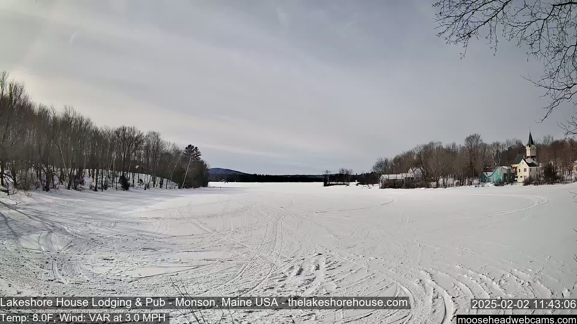 A frozen lake stretches out before a line of trees and houses under a mostly clear sky.