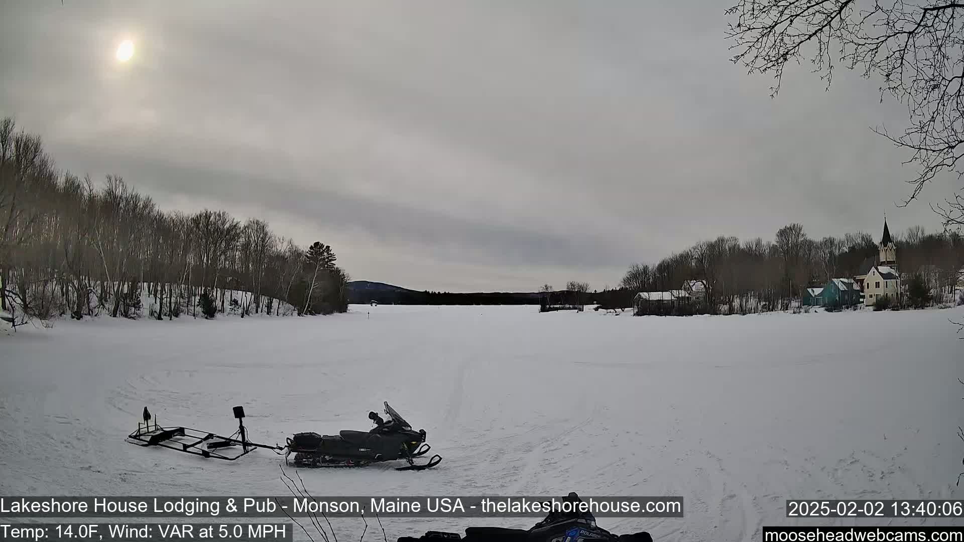 A snowmobile sits on a frozen lake under a cloudy sky, with a village visible in the distance.