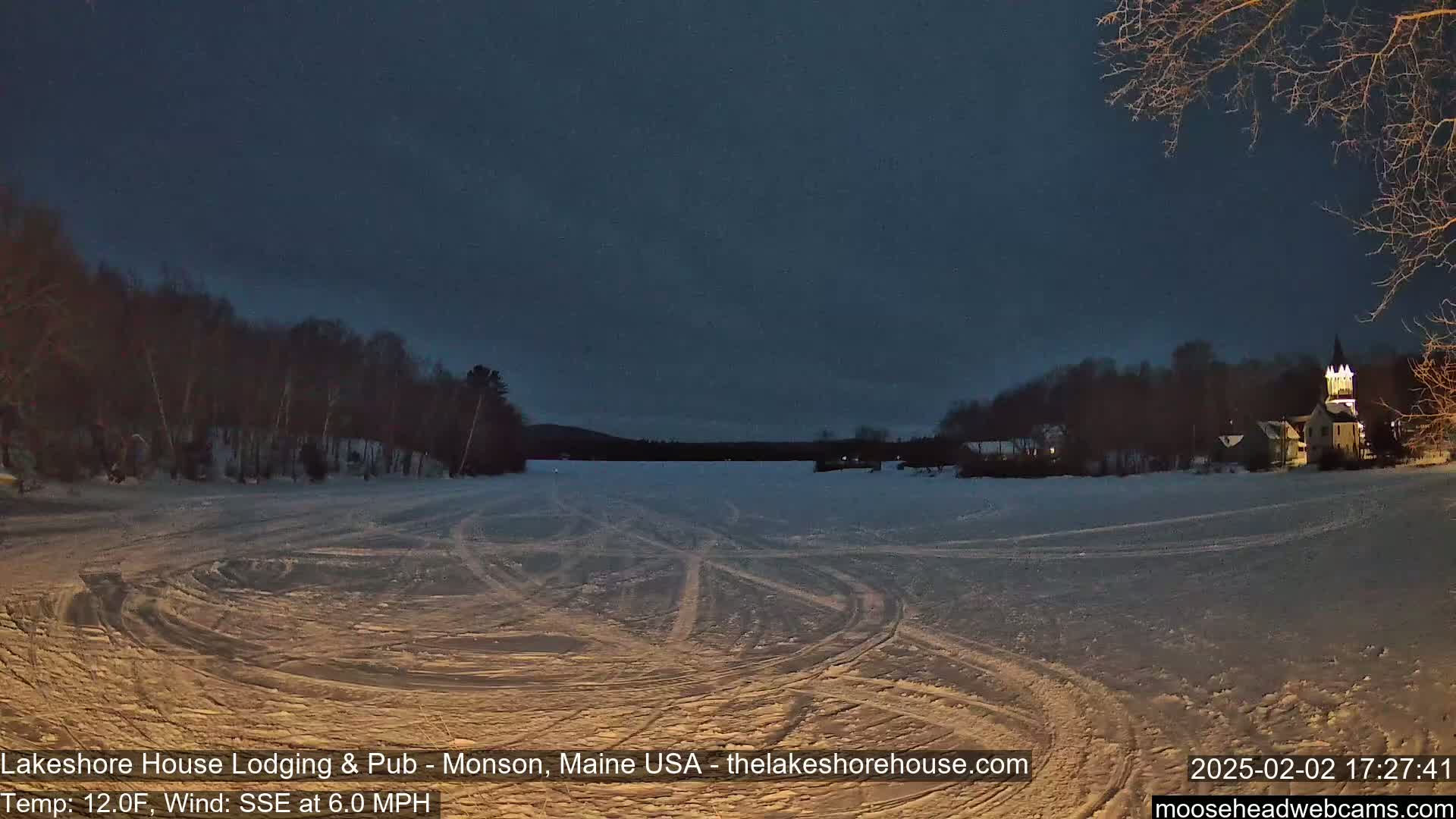 A snow-covered frozen lake at night, with a sparsely lit town and trees in the background under a dark sky.