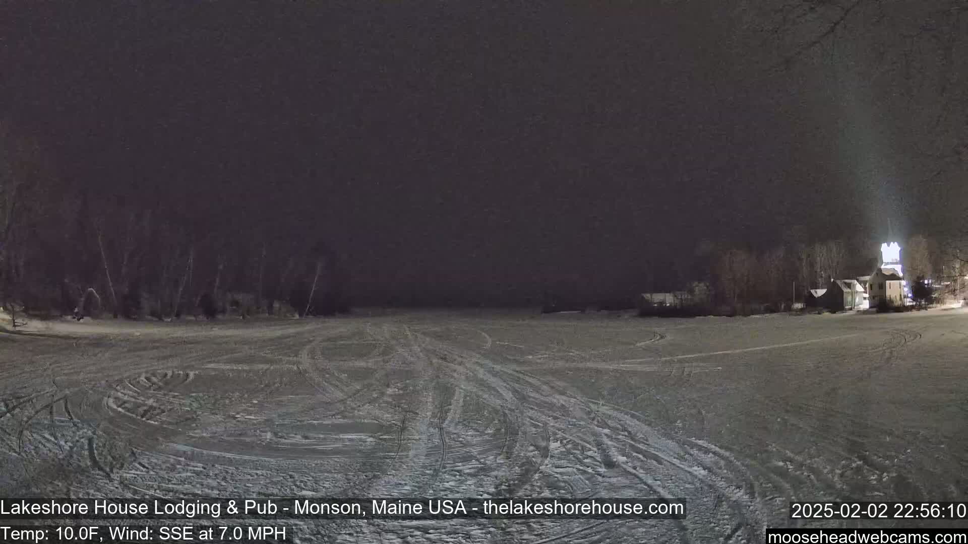 A snow-covered, flat expanse at night shows tire tracks, with dark trees and a lit building in the distance under a dark sky.