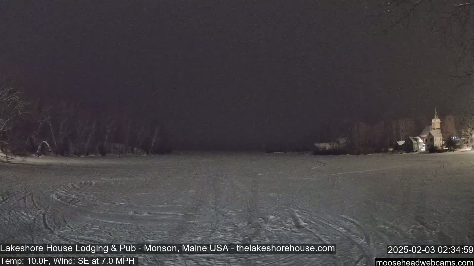 A snow-covered expanse stretches before a dark, nighttime scene featuring a building and trees under a dark sky.