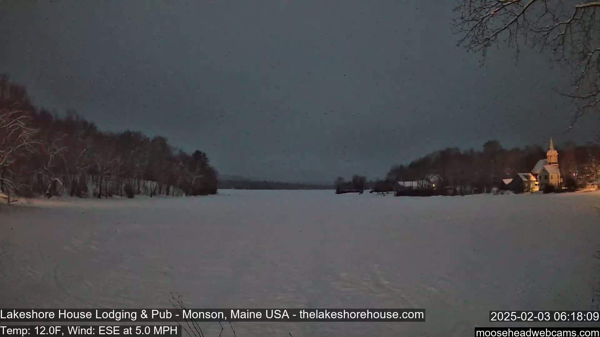 A snow-covered lake is seen at twilight, with a lightly snow-covered shoreline featuring buildings and trees under a dark, lightly snowing sky.