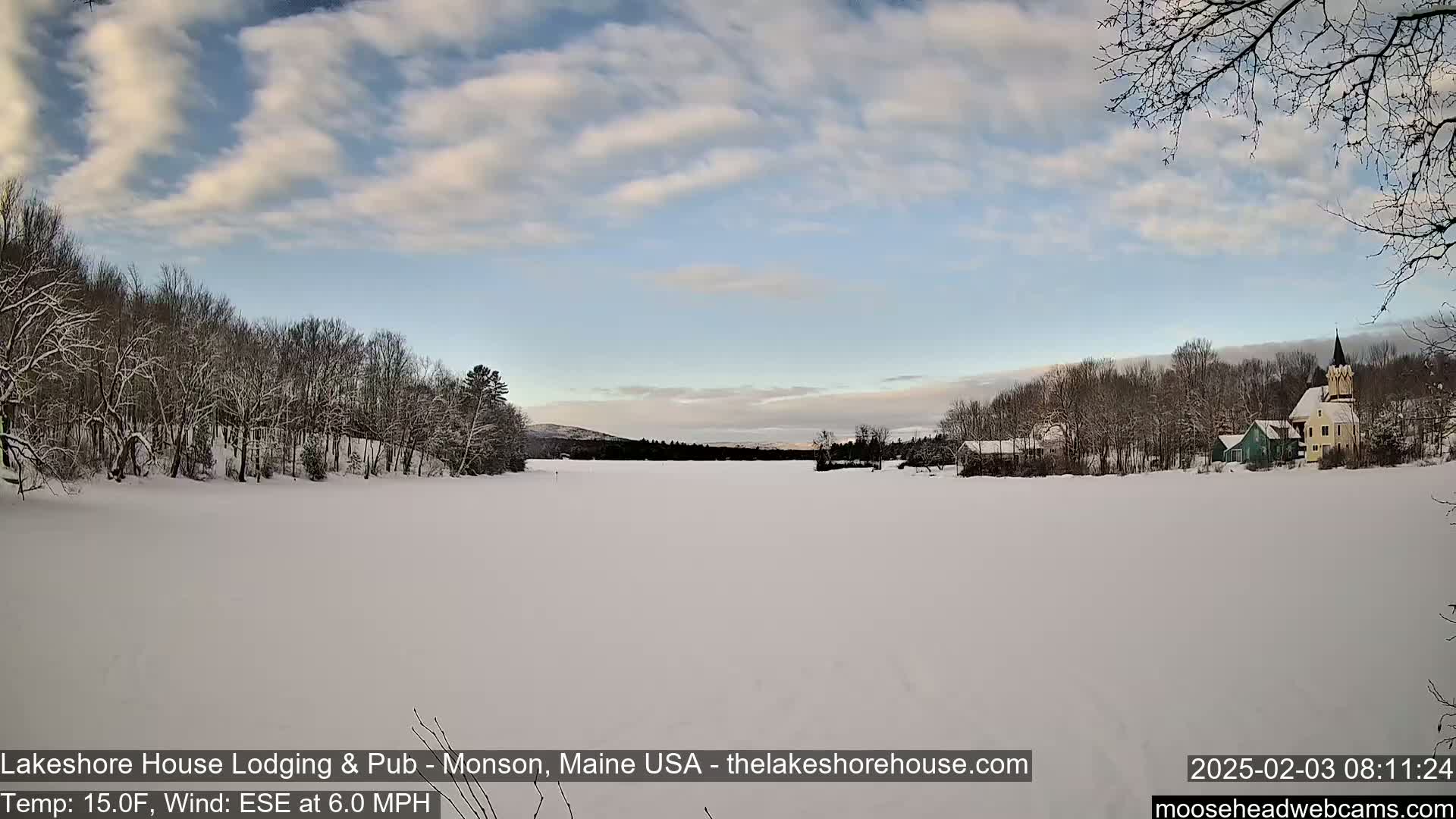 A snow-covered frozen lake stretches out before a line of snow-dusted trees and buildings under a partly cloudy sky.